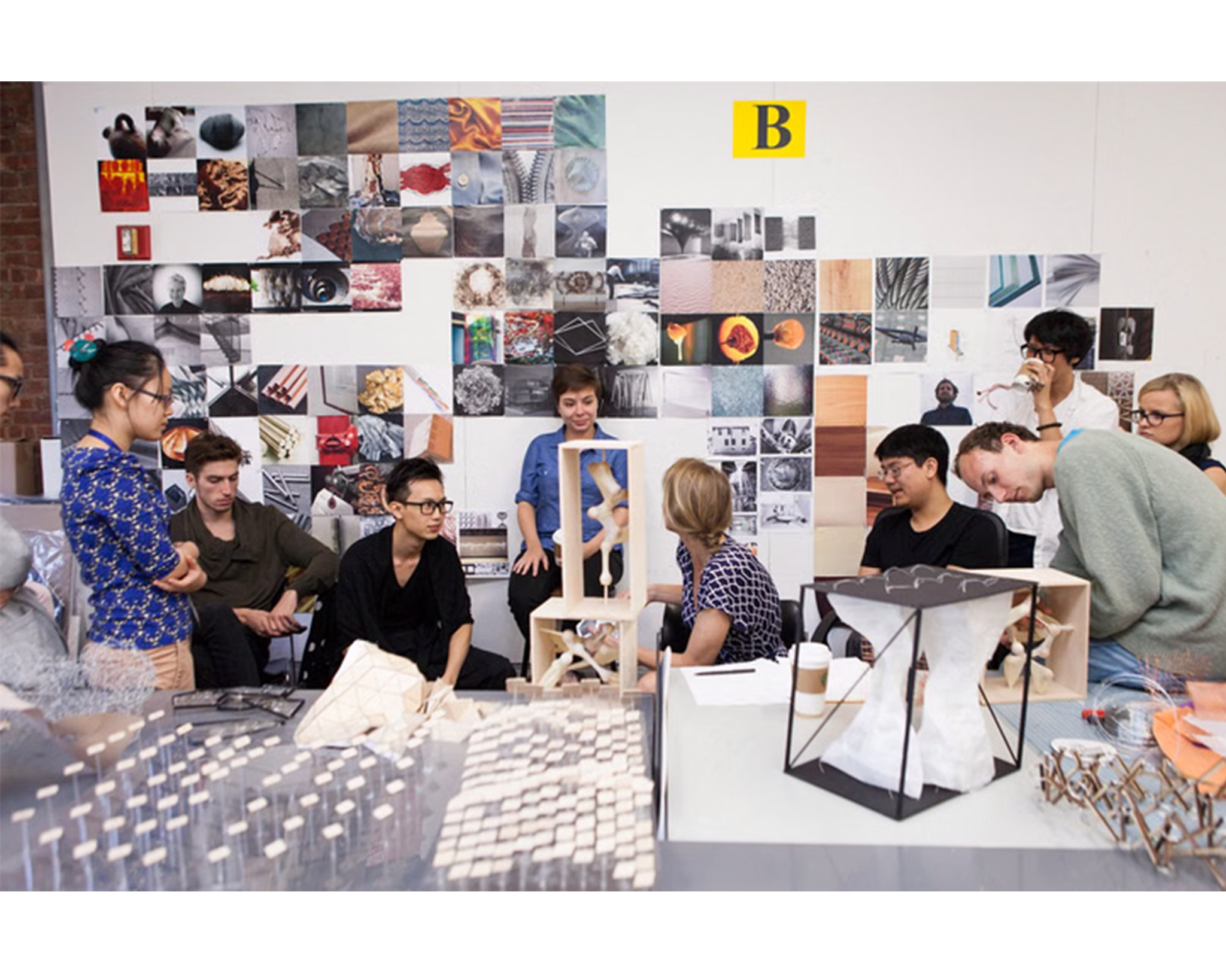 A group of students and faculty are gathered in a creative workspace discussing various architectural models. In the foreground, two intricate model designs are placed on a table filled with smaller components. Behind them, a collage of images adorns the wall, showcasing textures and abstract designs. Several participants are seated while others stand and engage in the conversation, displaying diverse expressions of interest and collaboration.