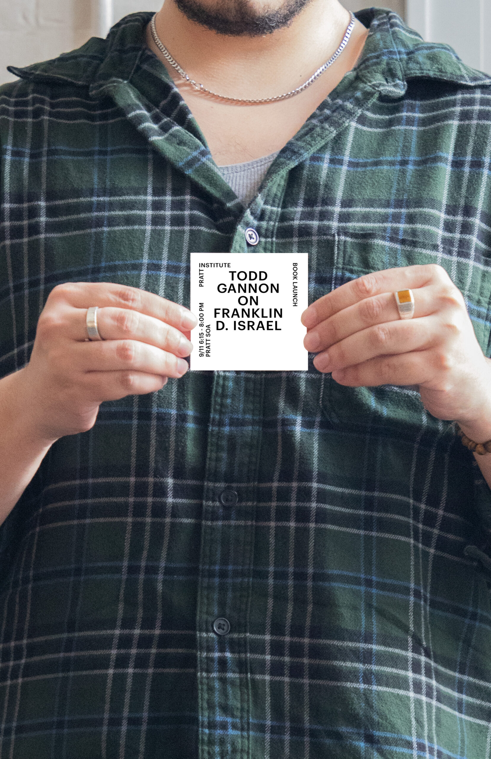 A person wearing a green plaid shirt holds a small printed card promoting “Todd Gannon on Franklin D. Israel,” a book launch event at Pratt Institute. The event details are displayed in clean black text on a white square.