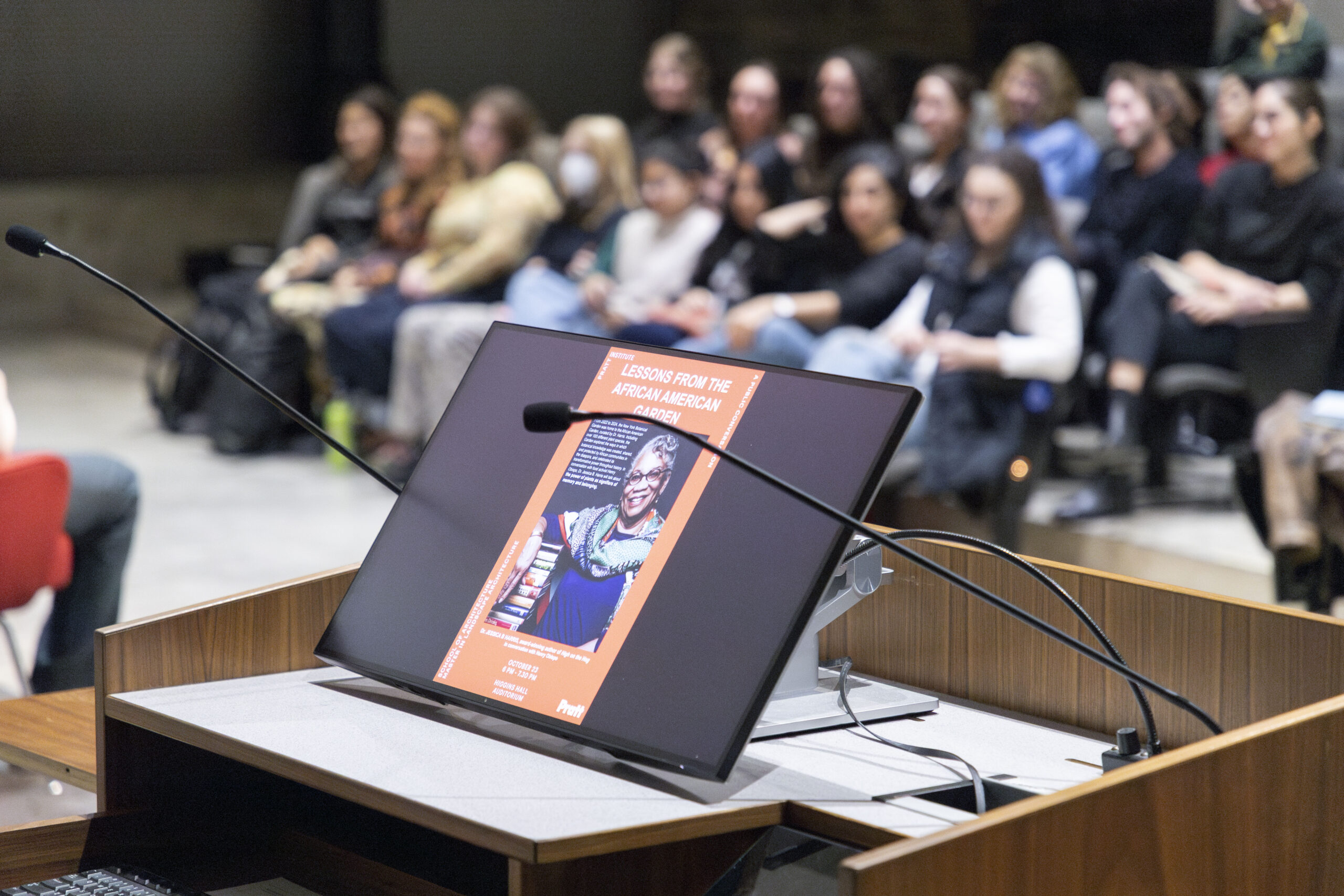 A presentation screen displaying the title "Lessons from the African American Garden," alongside an image of a woman, is positioned on a podium. A microphone is present in front of the screen. In the background, a seated audience of diverse individuals can be seen, engaging with the presentation.