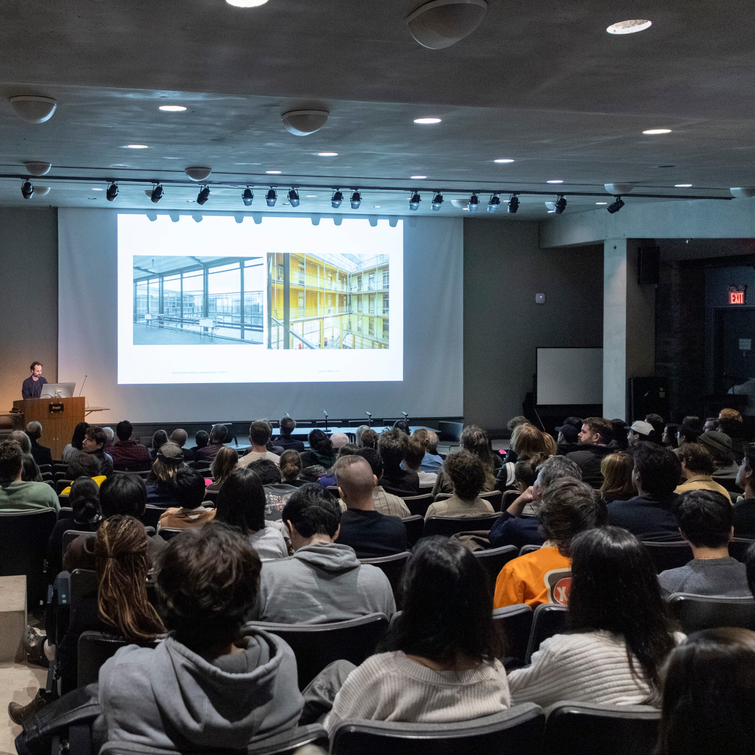 A speaker stands at a podium, presenting to an audience. The podium features a computer monitor displaying a graphic labeled "HIGHWAY" with a simple drawing. The setting is an indoor auditorium with a table visible in the foreground, holding water bottles. The lighting is soft, highlighting the speaker and the presentation backdrop.