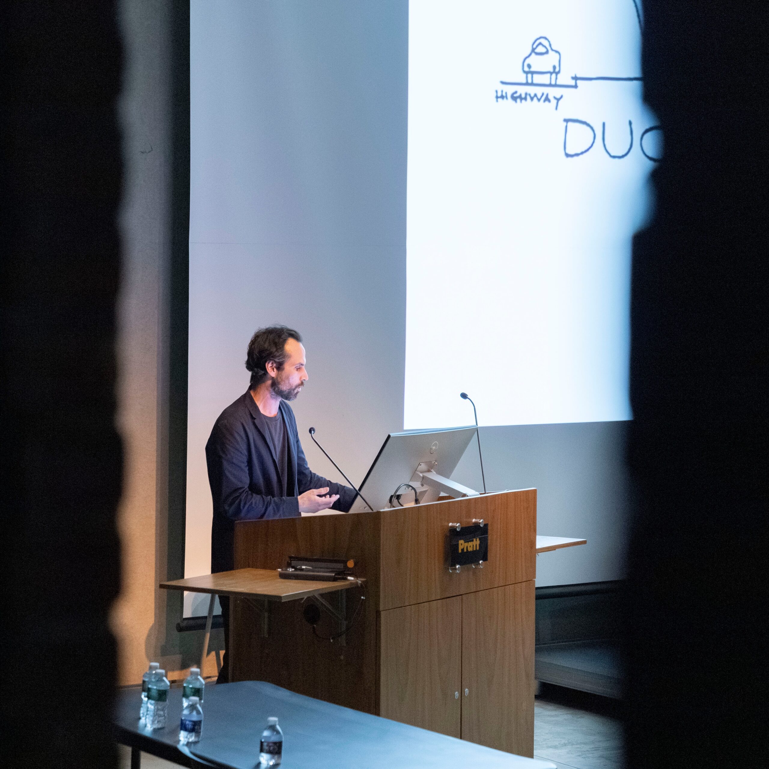 A man with short, dark hair is standing at a wooden podium, presenting during a lecture. He is wearing a dark sweater and appears to be using hand gestures to emphasize his points. A screen behind him displays a diagram labeled "HIGHWAY" and "DUC." There are water bottles on the table in front of him, and part of the audience is visible in the shadows on either side.