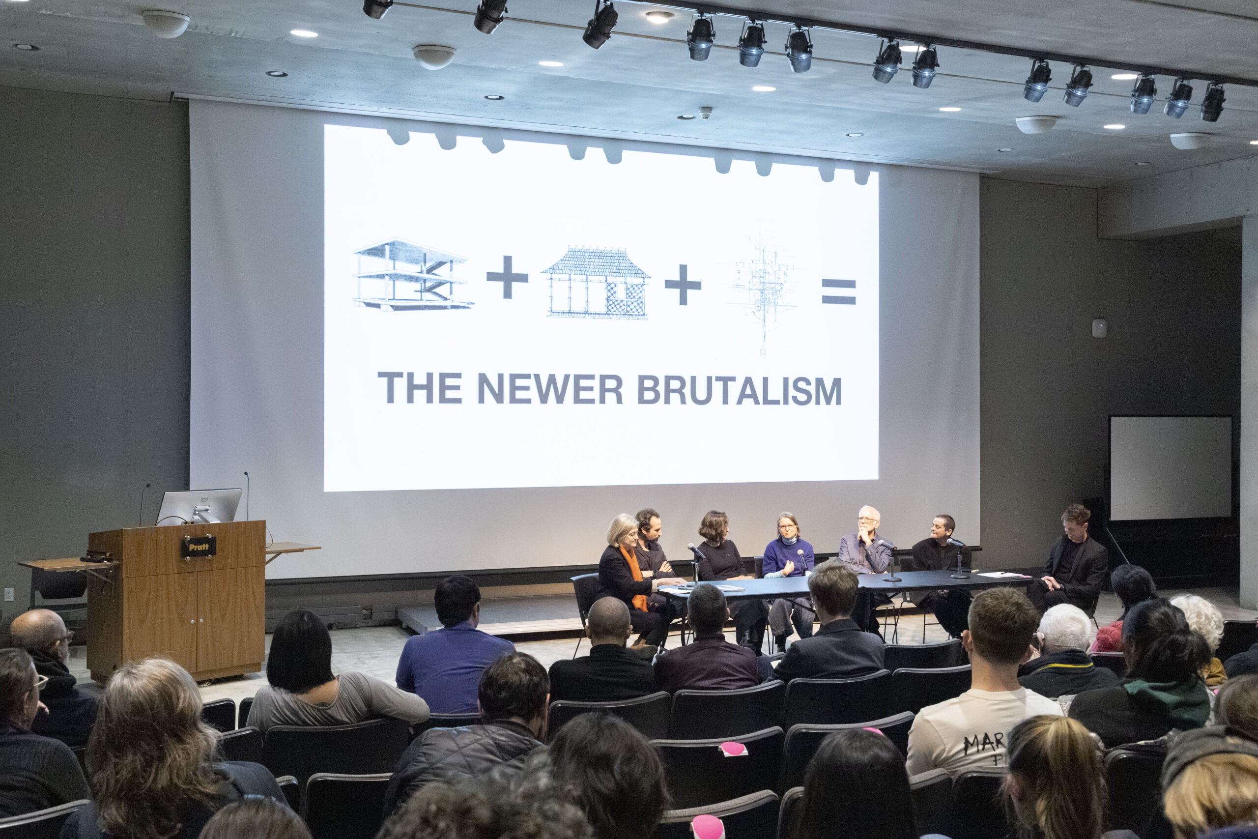 A man with dark hair and a beard stands at a podium during a presentation. He is facing the audience and gesturing towards a large screen displaying a diagram, including the text "HIGHWAY" and "DUC." The setting appears to be an auditorium, with a table in front holding water bottles and a dark surface.