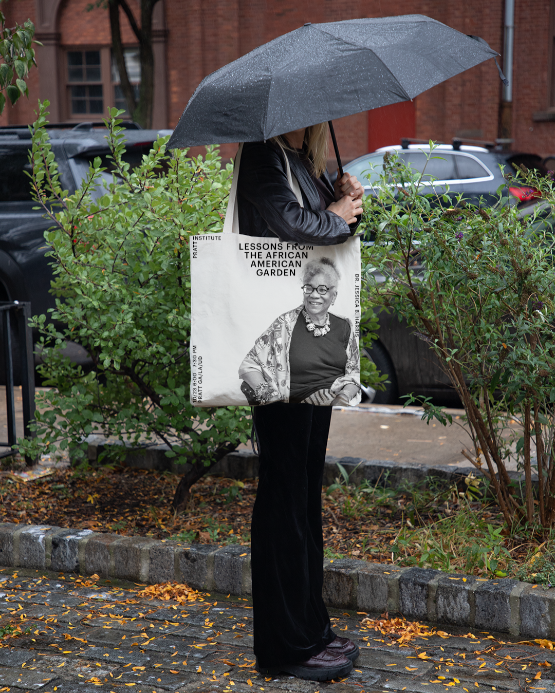 A person wearing a black jacket is holding a tote bag. The bag features a black and white photo of a smiling older woman wearing glasses, adorned with jewelry and a colorful shawl. Text on the bag states "LESSONS FROM THE AFRICAN AMERICAN GARDEN" and "DR. JESSICA B. HARRIS," along with event details: "10/23 6:00 - 7:30 PM, PRATT GALA/UD." The background shows green foliage.