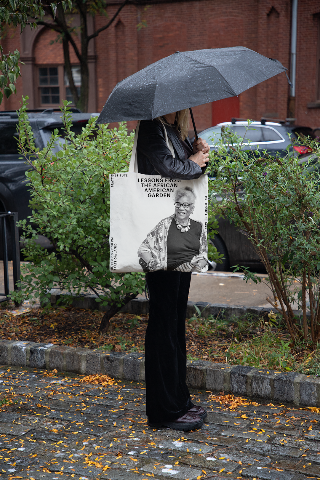 A person wearing a black jacket is holding a tote bag. The bag features a black and white photo of a smiling older woman wearing glasses, adorned with jewelry and a colorful shawl. Text on the bag states "LESSONS FROM THE AFRICAN AMERICAN GARDEN" and "DR. JESSICA B. HARRIS," along with event details: "10/23 6:00 - 7:30 PM, PRATT GALA/UD." The background shows green foliage.