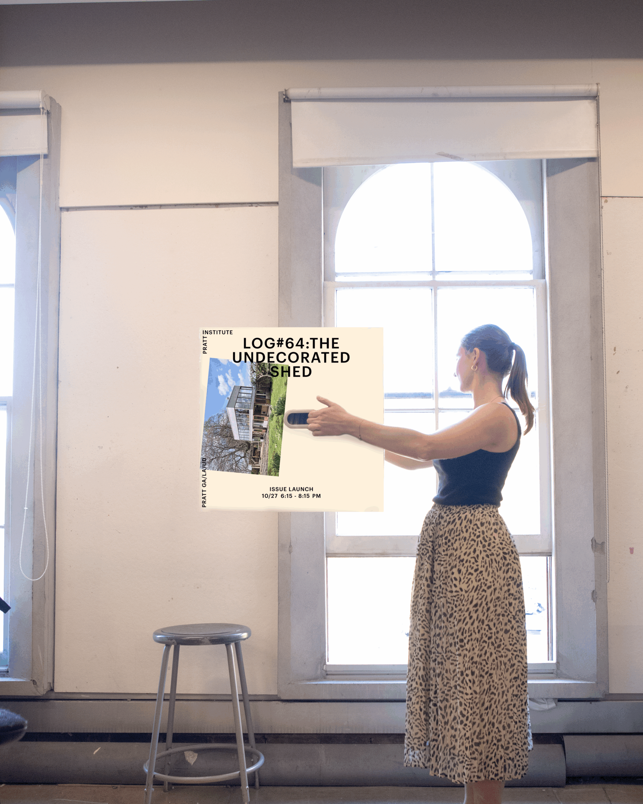 A woman stands in a room holding a poster titled "LOG#64: THE UNDECORATED SHED." She wears a black tank top and a long, patterned skirt. Behind her, large windows let in natural light. A stool is positioned in front of her. The poster includes details about an issue launch event.