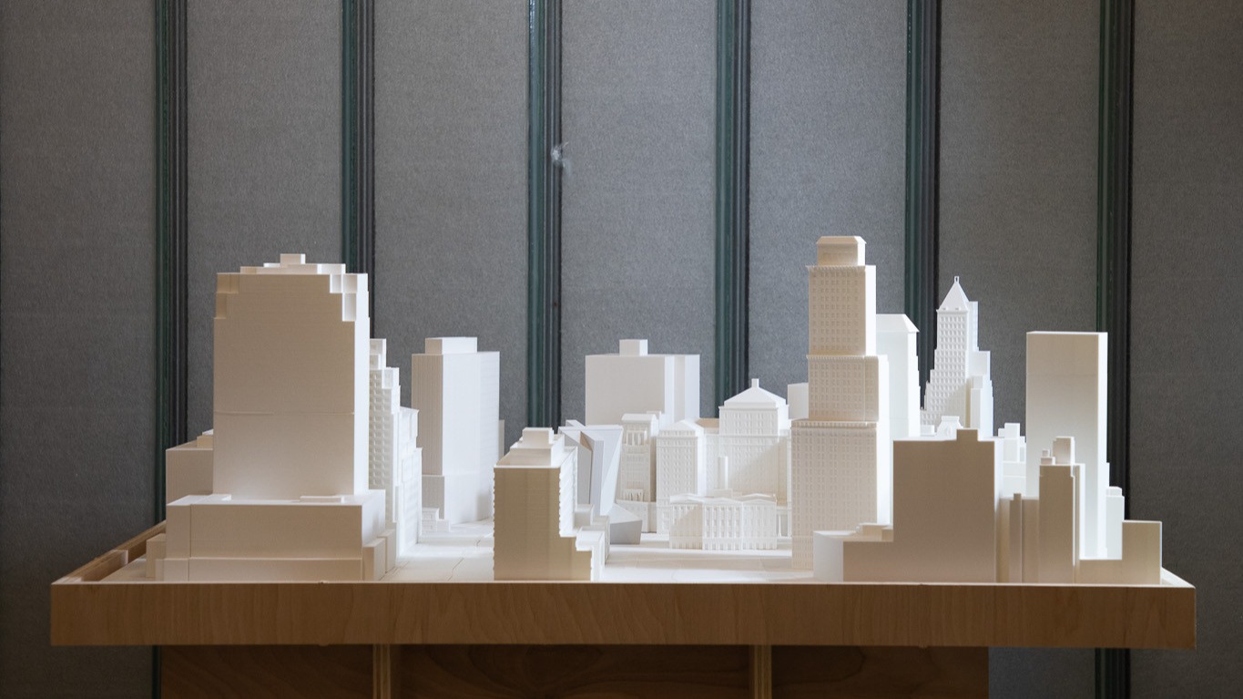 A white architectural model of a cityscape displayed on a wooden table. The model features various building heights and shapes, showcasing a skyline with detailed structures. The background has a neutral gray fabric, adding contrast to the model.