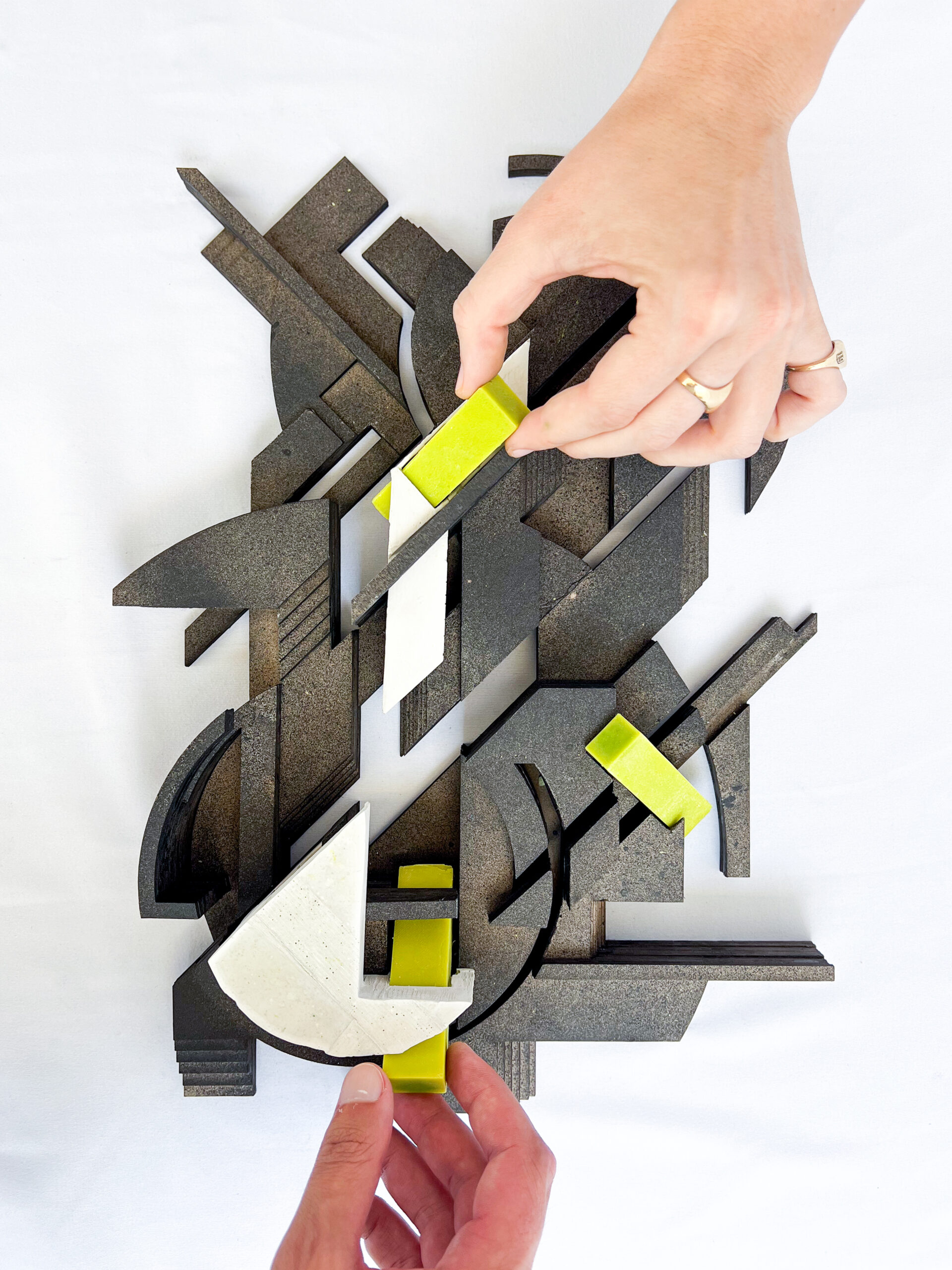 Two hands are interacting with an abstract arrangement of geometric shapes in a mix of black and white materials, along with bright yellow accents. The composition features a variety of textures and forms, showcasing a creative and tactile exploration. The background is a plain white surface, enhancing the contrast between the elements.
