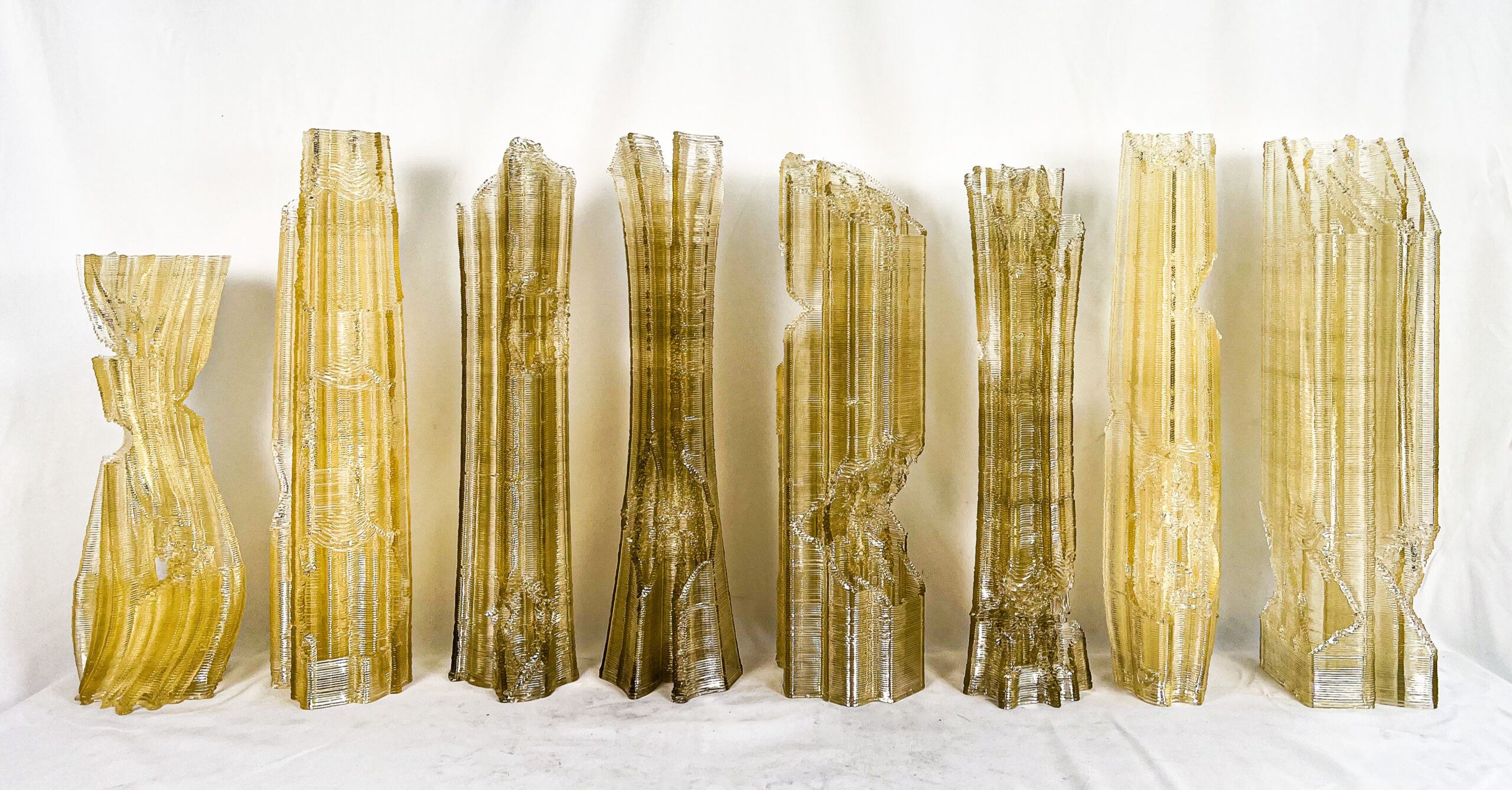 A collection of seven tall, sculptural pieces made from a yellowish translucent material. Each piece has a distinct shape and texture, resembling tall, textured columns with jagged edges and layered patterns. They are arranged in a line against a light background, creating an artistic display that emphasizes their unique forms and surfaces.