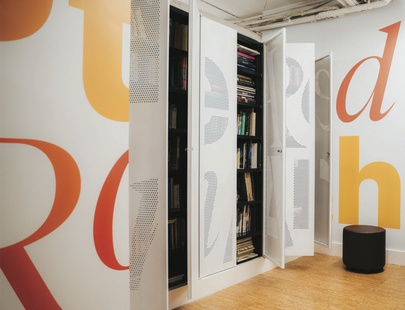 A modern interior with large, colorful letters on the wall. Two white cabinets with perforated designs are open, revealing shelves filled with books. A small, round black stool is situated on the floor, which is made of light-colored bamboo or wood. The overall atmosphere appears to be a creative or study space.