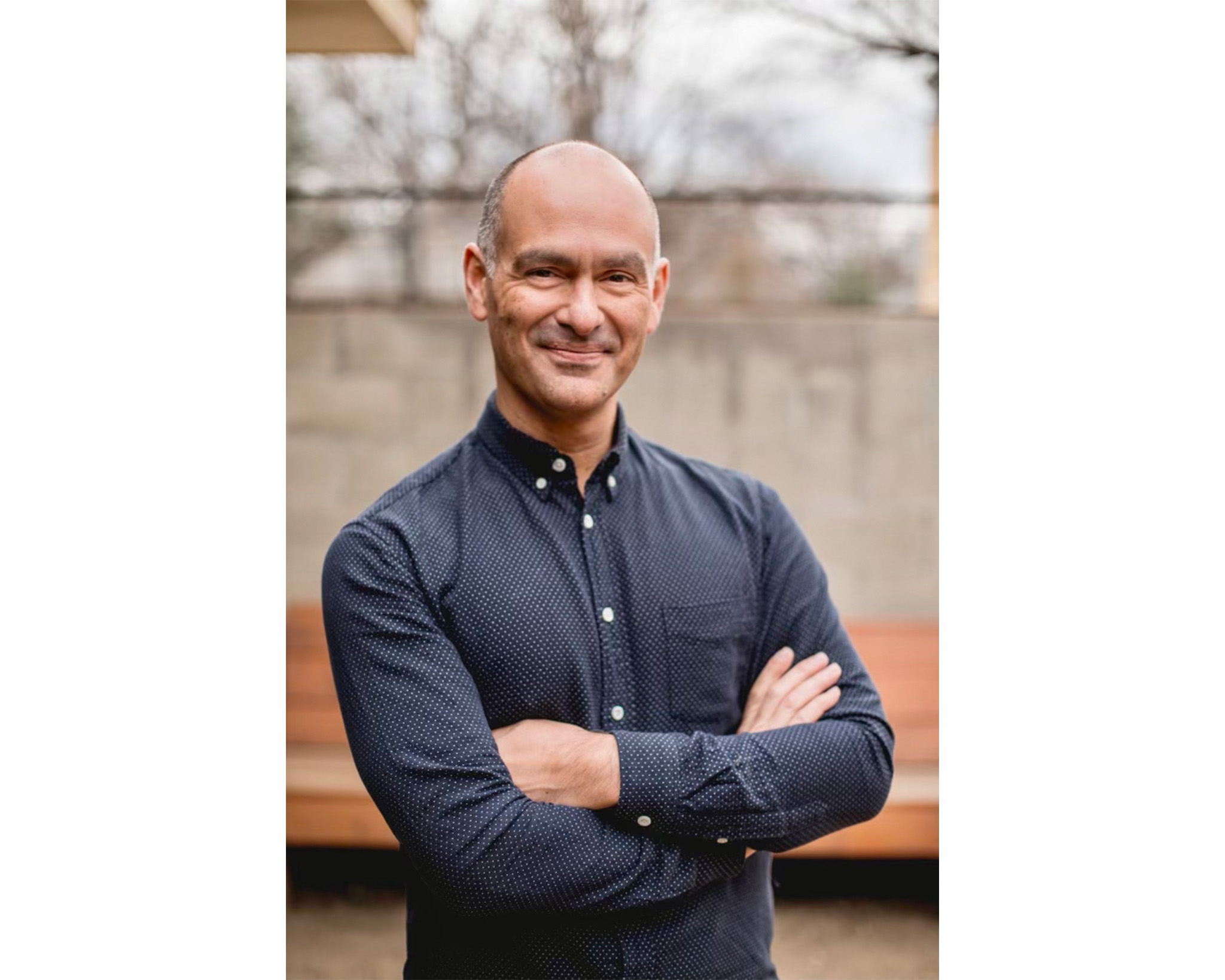 A smiling man stands outdoors, arms crossed, wearing a dark blue, long-sleeved button-up shirt with small polka dots. He has short, shaved hair and is positioned against a blurred background of trees and wooden benches, suggesting a relaxed environment.