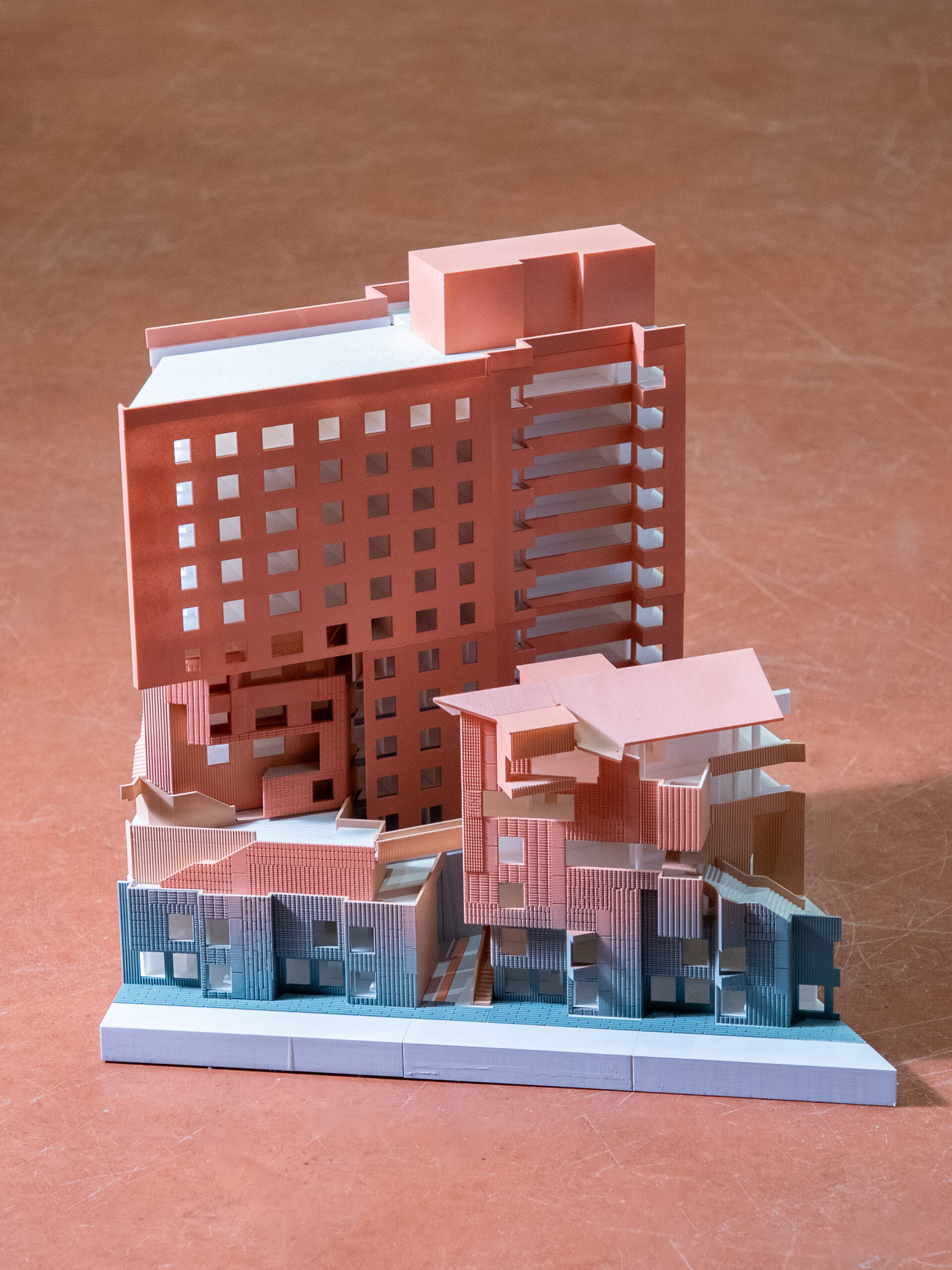 A detailed architectural model featuring multiple buildings. The structures are designed with various geometric shapes, layered forms, and a mix of coral and blue colors. The model shows a blend of flat and textured surfaces, with some transparent elements. It is presented on a white base, set against a warm-toned floor.