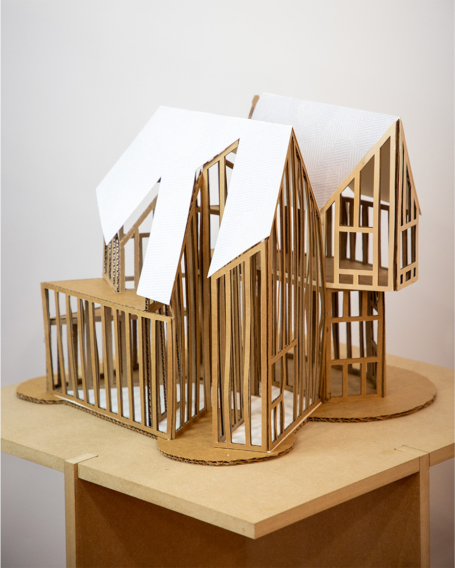 A model of a building constructed from cardboard, featuring a frame-like design with exposed structural elements. The model has two distinct structures with sloped roofs covered in textured white paper. It is displayed on a circular cardboard base atop a wooden table, against a plain white wall.