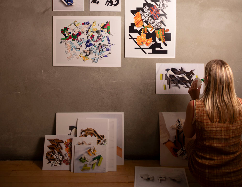 A person with long hair is standing in front of a wall displaying various colorful artworks, composed of cut-out shapes and designs. The individual appears to be attaching or adjusting one of the artworks, using tape or adhesive. Several framed images are propped against the wall and stacked on the floor, creating a creative and artistic environment. The background is a muted gray, contrasting with the vibrant colors of the artworks.