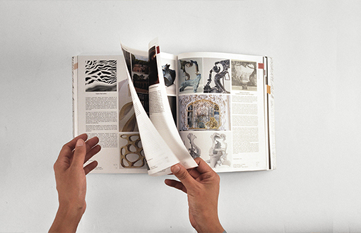 A pair of hands is shown flipping through the pages of a large open book. The pages contain various artworks and illustrations alongside descriptive text. The background is plain and white, emphasizing the book and hands.