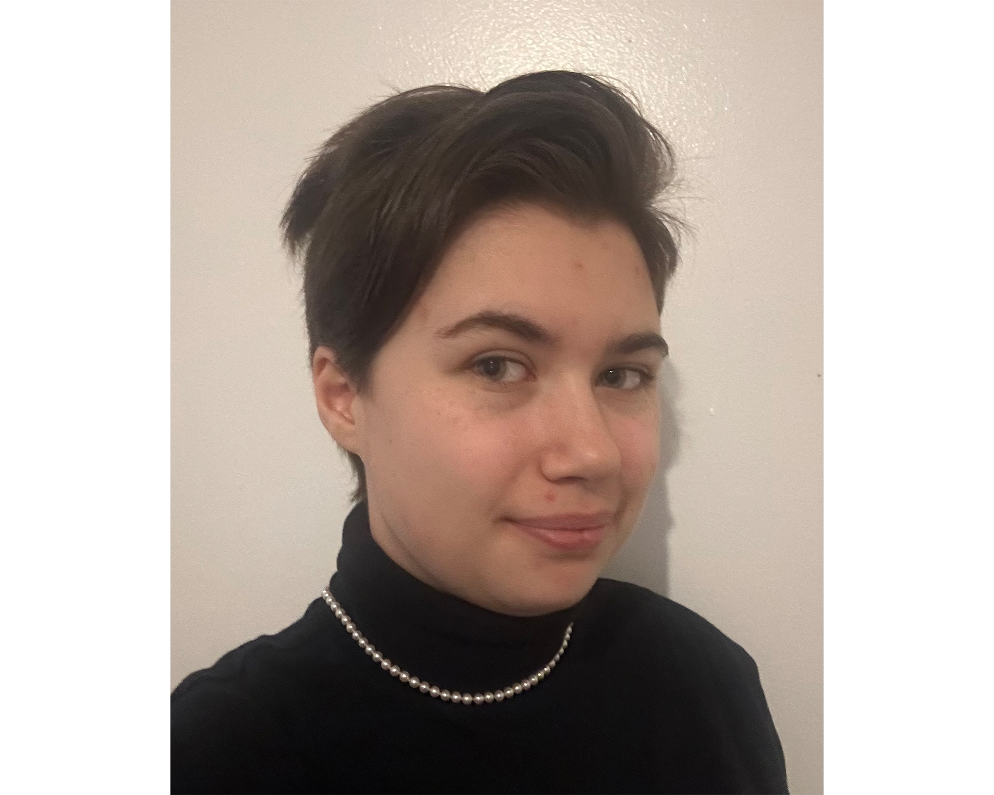 A close-up portrait of a person with short, dark hair, wearing a black turtleneck sweater and a pearl necklace. The background is a plain, light-colored wall. The person is smiling slightly and facing slightly to the side.