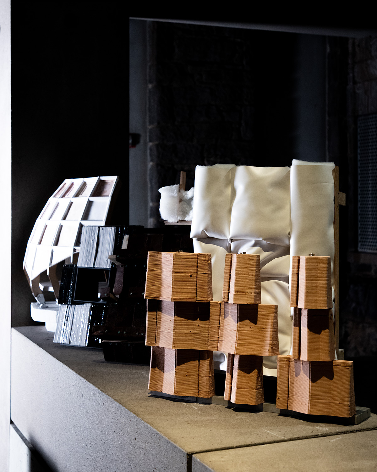 A collection of architectural models is displayed on a concrete ledge. The models vary in material and form, with some made of smooth, white fabric and others featuring textured, earthy-toned elements that resemble clay or terracotta. The setting is dimly lit, which accentuates the textures and shapes of the models, creating an artistic and experimental atmosphere.
