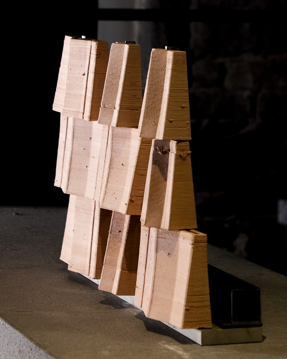 A sculptural arrangement of several stacked, rectangular wooden blocks, varying in height and positioned at different angles. The wood has a natural finish with visible grain patterns and small imperfections. The blocks are mounted on a flat surface, contrasting against a dark background. The overall composition creates a dynamic visual effect.