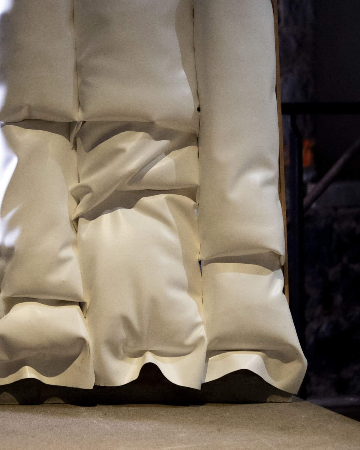 A close-up view of a series of large, white, padded sections, possibly made of a soft material. The sections are arranged in a stacked formation, with creases and folds indicating their softness. The bottom edge is slightly curved, revealing a contrasting dark or textured surface beneath. The background appears blurred, suggesting an indoor setting.