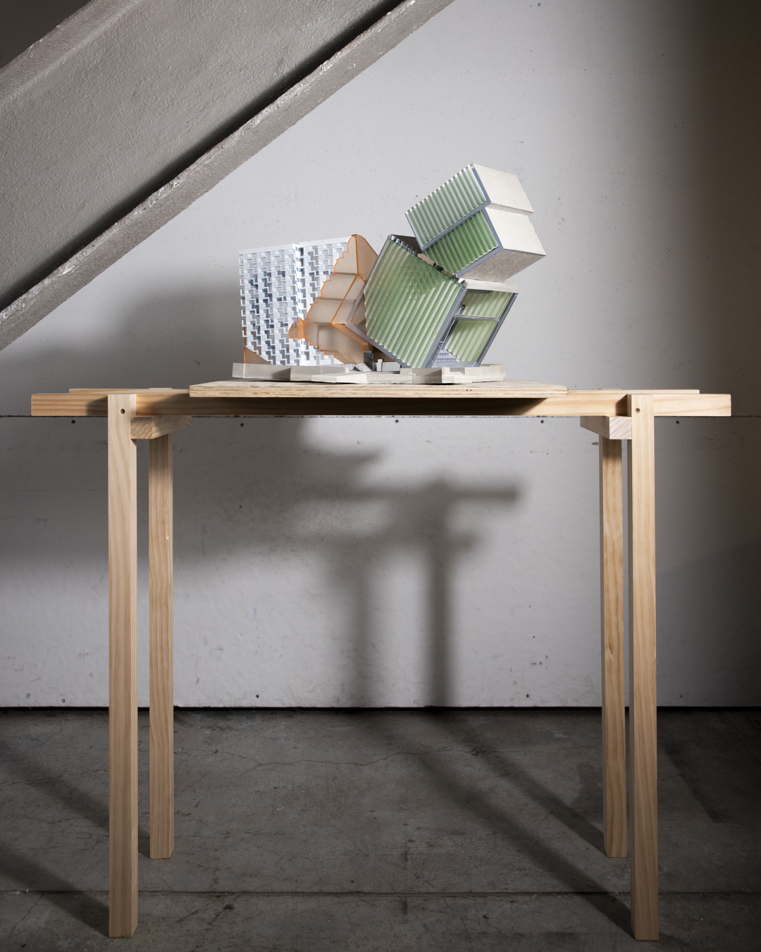 A wooden table with four legs supports a sculptural model composed of several geometric shapes representing buildings. The structures, featuring various textures and colors, appear to be precariously balanced. The background includes a concrete wall and part of a staircase, creating a minimalistic studio environment.