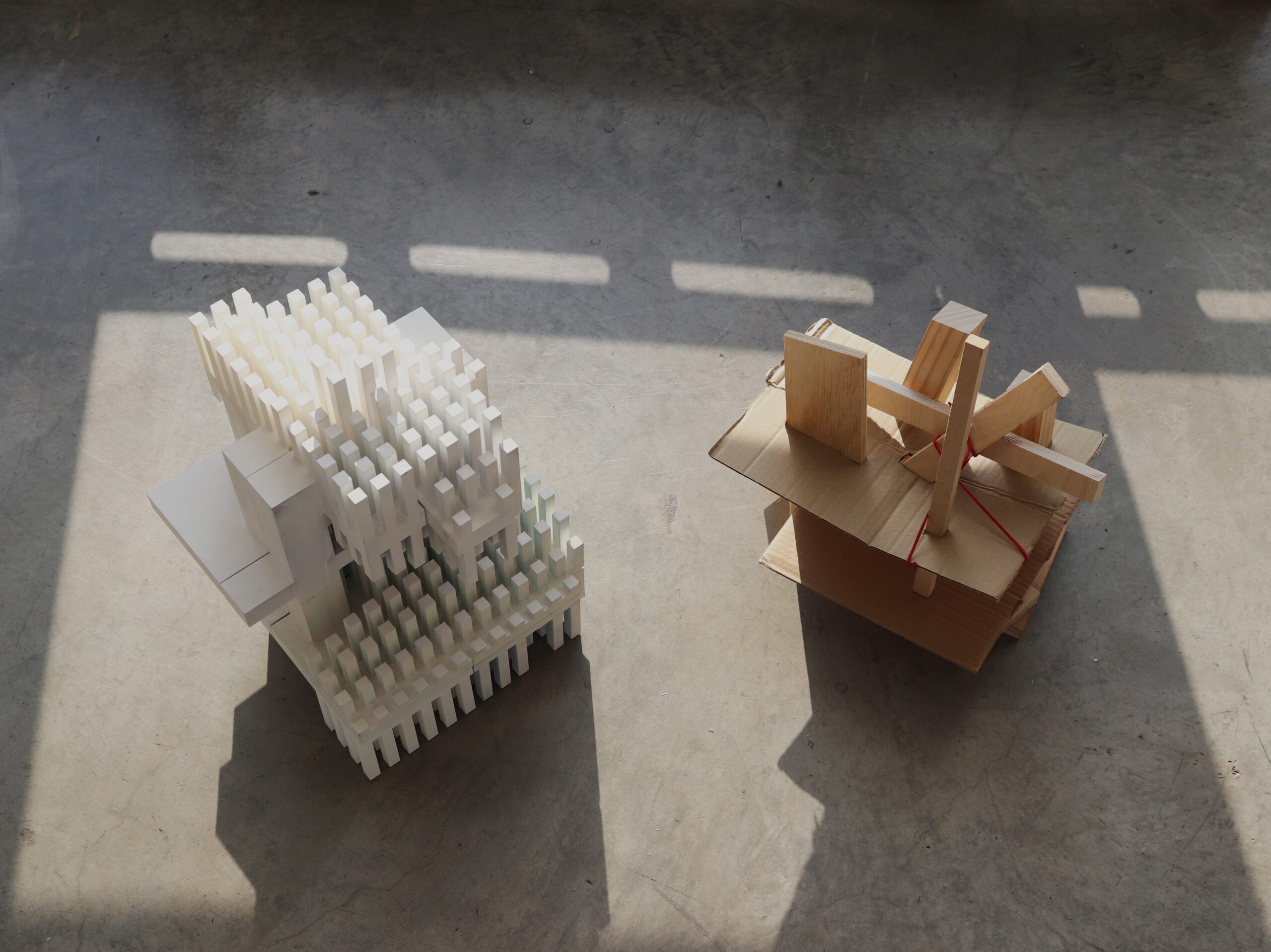 Two architectural models are displayed on a concrete surface. The model on the left is constructed from white materials, featuring stacked rectangular forms with vertical elements. The model on the right is made of wood and cardboard, with a more abstract arrangement of wooden pieces and a simple base. Sunlight creates distinct shadows on the ground, enhancing the appearance of both models.