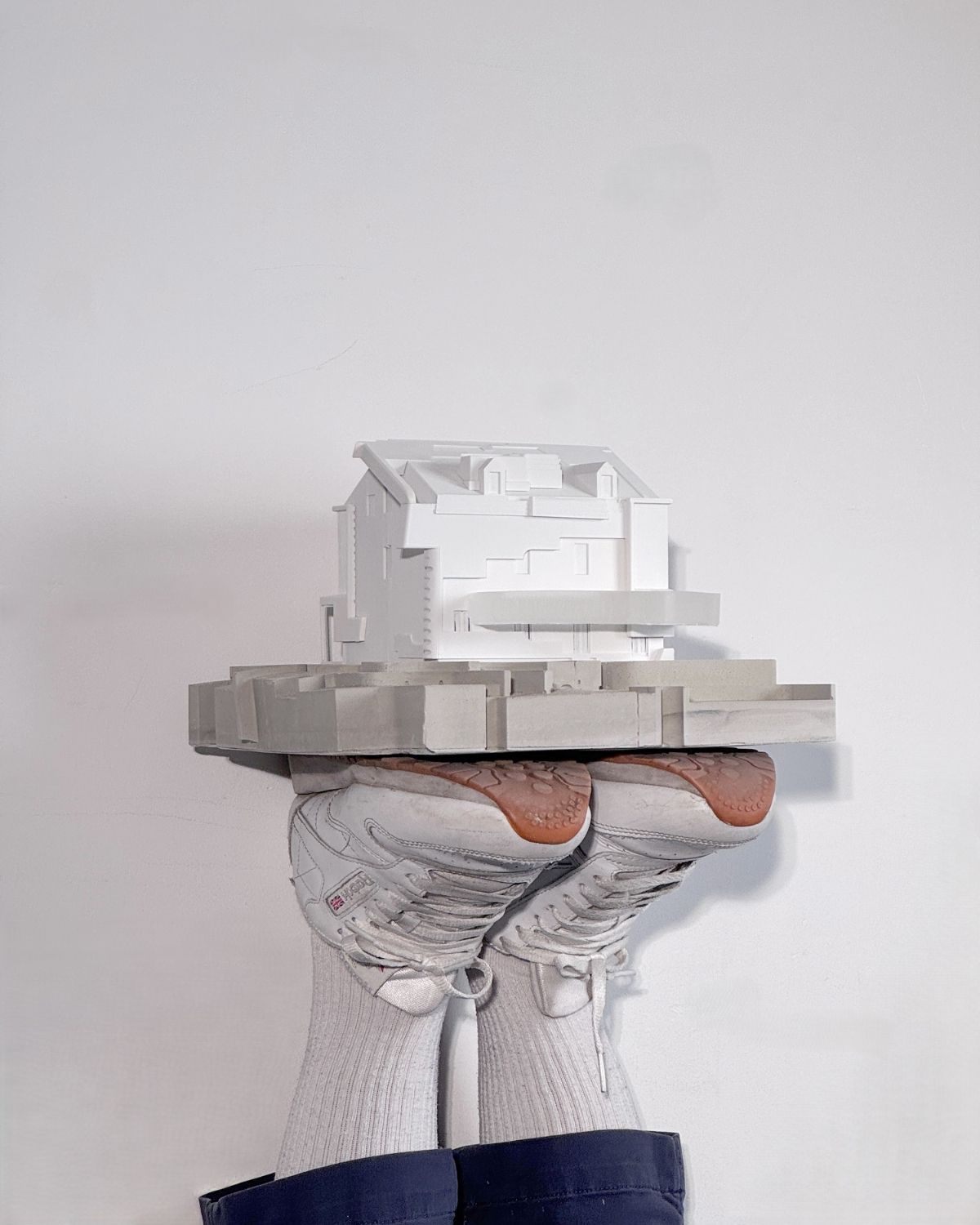 A pair of feet wearing white socks and sneakers supports a model of a white building. The building model is elevated above the feet, with a minimalistic design, featuring various geometric shapes and flat surfaces. The background is a plain white wall.