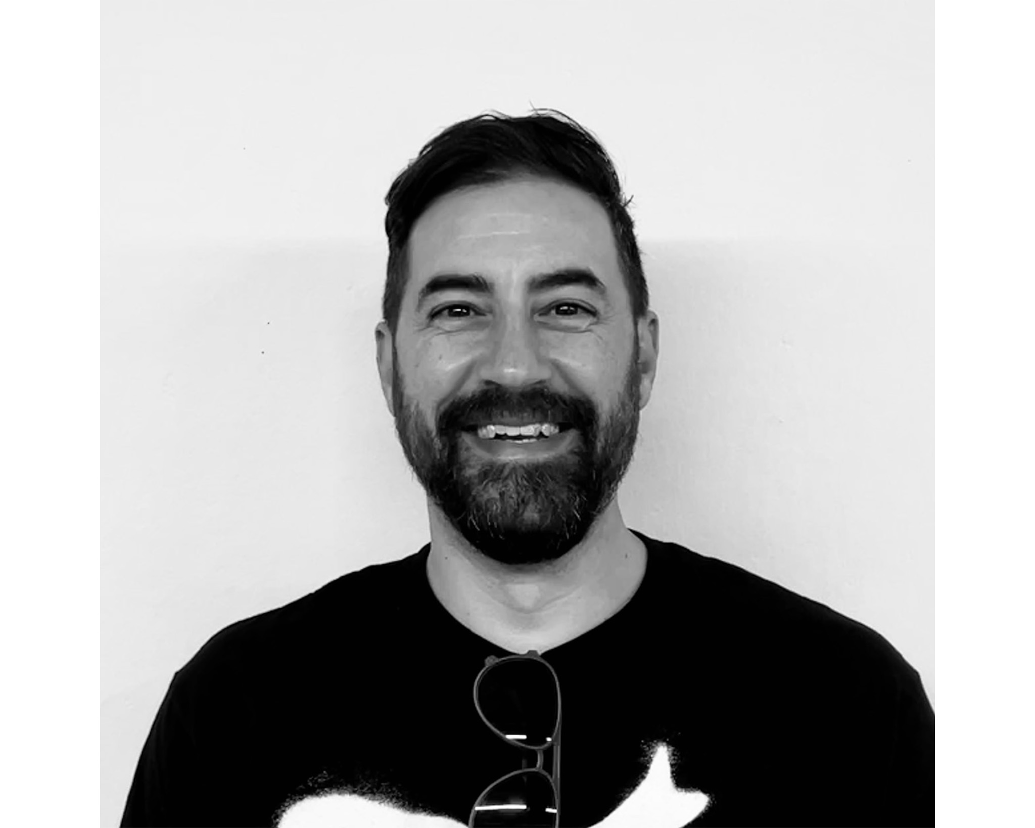 A smiling man with a beard and short, styled hair is facing the camera. He is wearing a black t-shirt with a white graphic. He has sunglasses resting on his chest, and the background is a plain white wall.