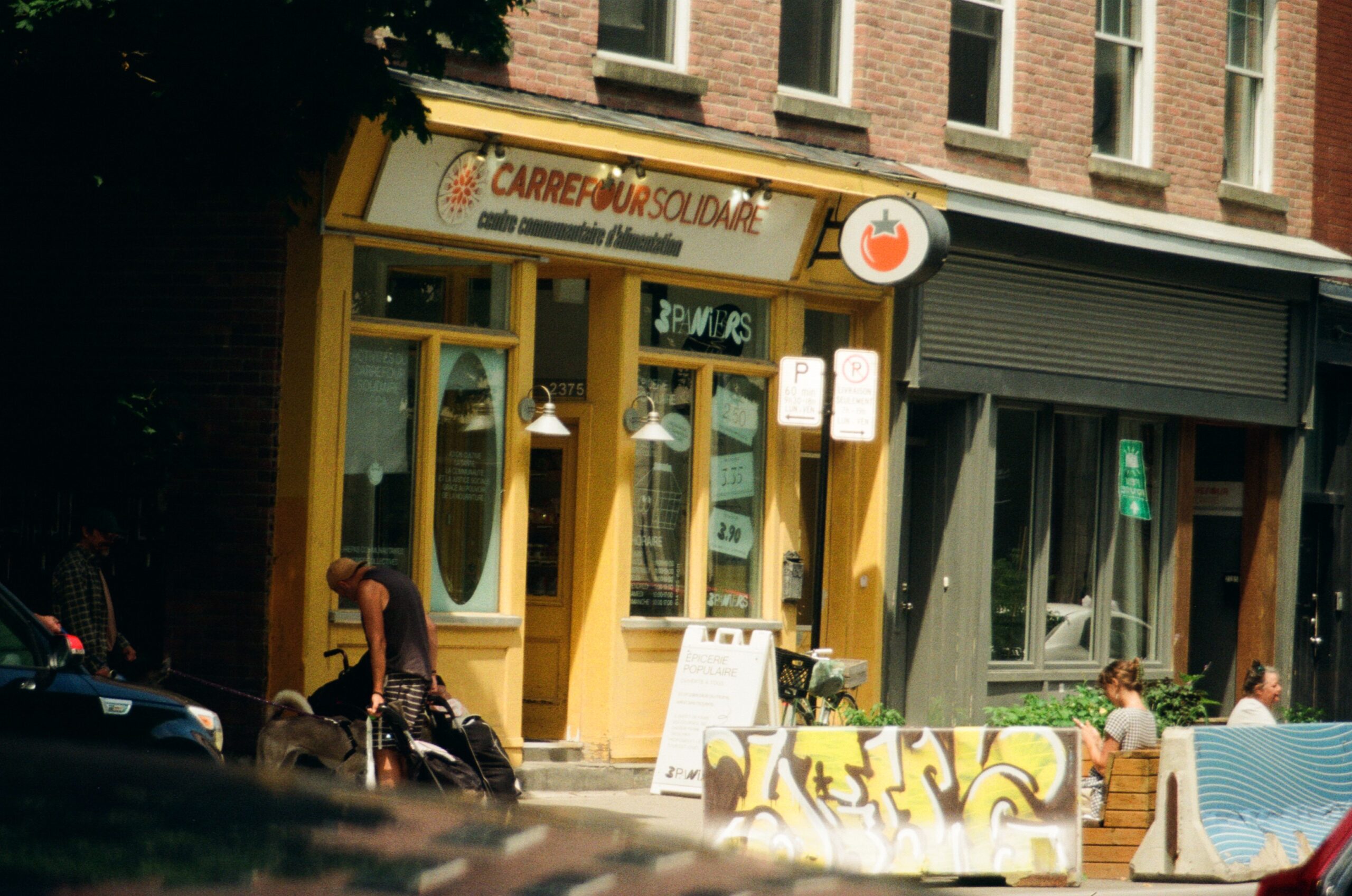 A vibrant community center called "Carrefour Solidaire" is featured in the image, showcasing a bright yellow exterior with large windows and a red tomato logo. In front of the center, two people interact, one with a dog on a leash, while another sits nearby. A sign with menu items and prices is displayed outside. The background includes a mix of brick buildings and a sidewalk with graffiti art on benches.