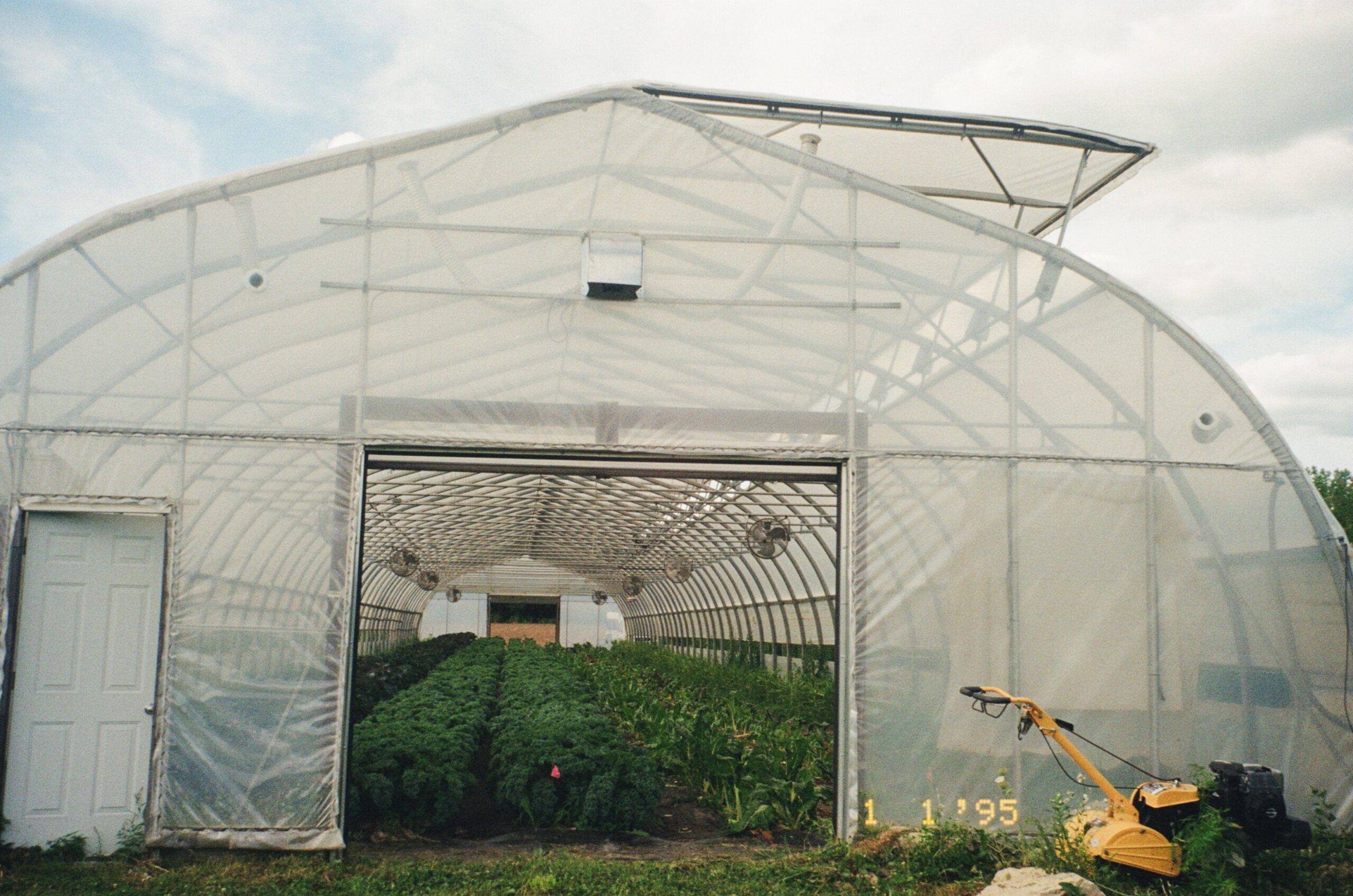 A large, translucent greenhouse with a white door on the left. Inside, rows of green plants are visible. The structure features ventilation fans along the sides and has a grassy area in front. A yellow gardening machine is parked nearby. The sky is partly cloudy, indicating fair weather.