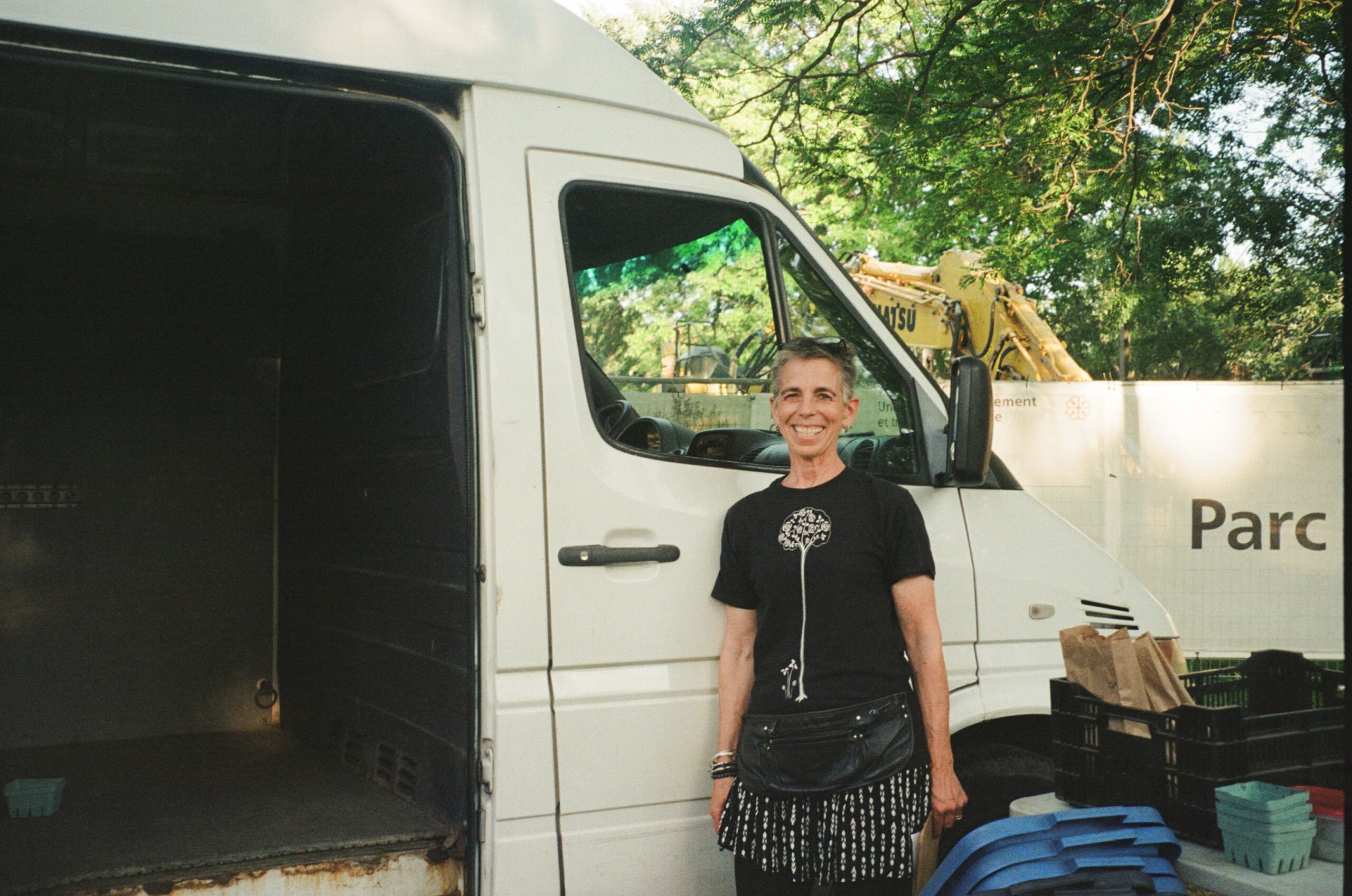 A smiling person stands beside a white van parked under trees. They wear a black t-shirt with a graphic design and a black bag with fringes. The van's door is open, revealing an empty cargo area, and there are boxes and baskets in various colors on the ground nearby. A construction site is visible in the background with a "Parc" sign.
