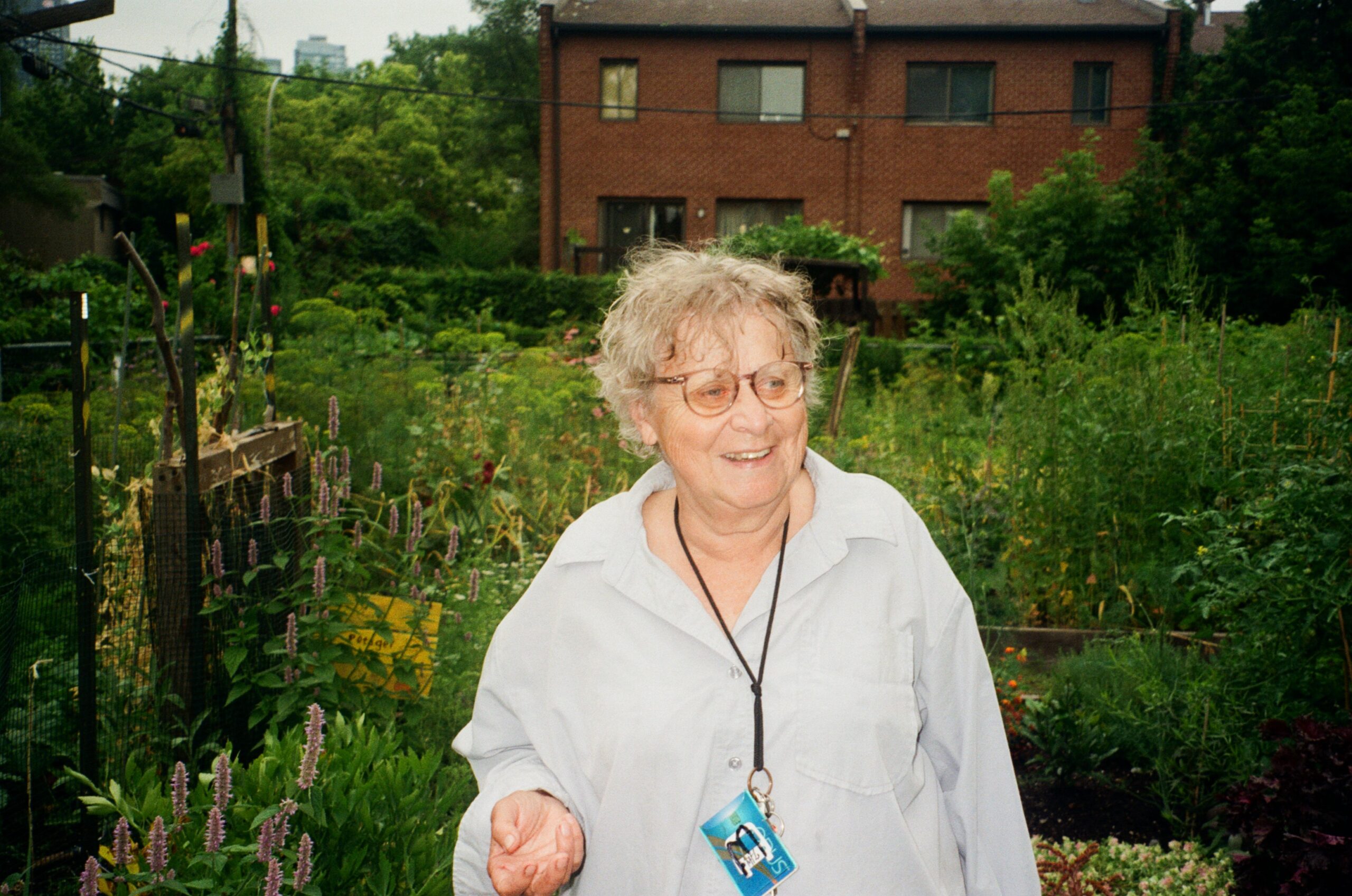 An elderly person with curly gray hair and glasses stands in a lush garden filled with various plants. They are wearing a light-colored button-up shirt and a lanyard with an ID attached. A brick building is visible in the background, partially obscured by greenery. The person has a friendly smile and appears to be enjoying the garden surroundings.