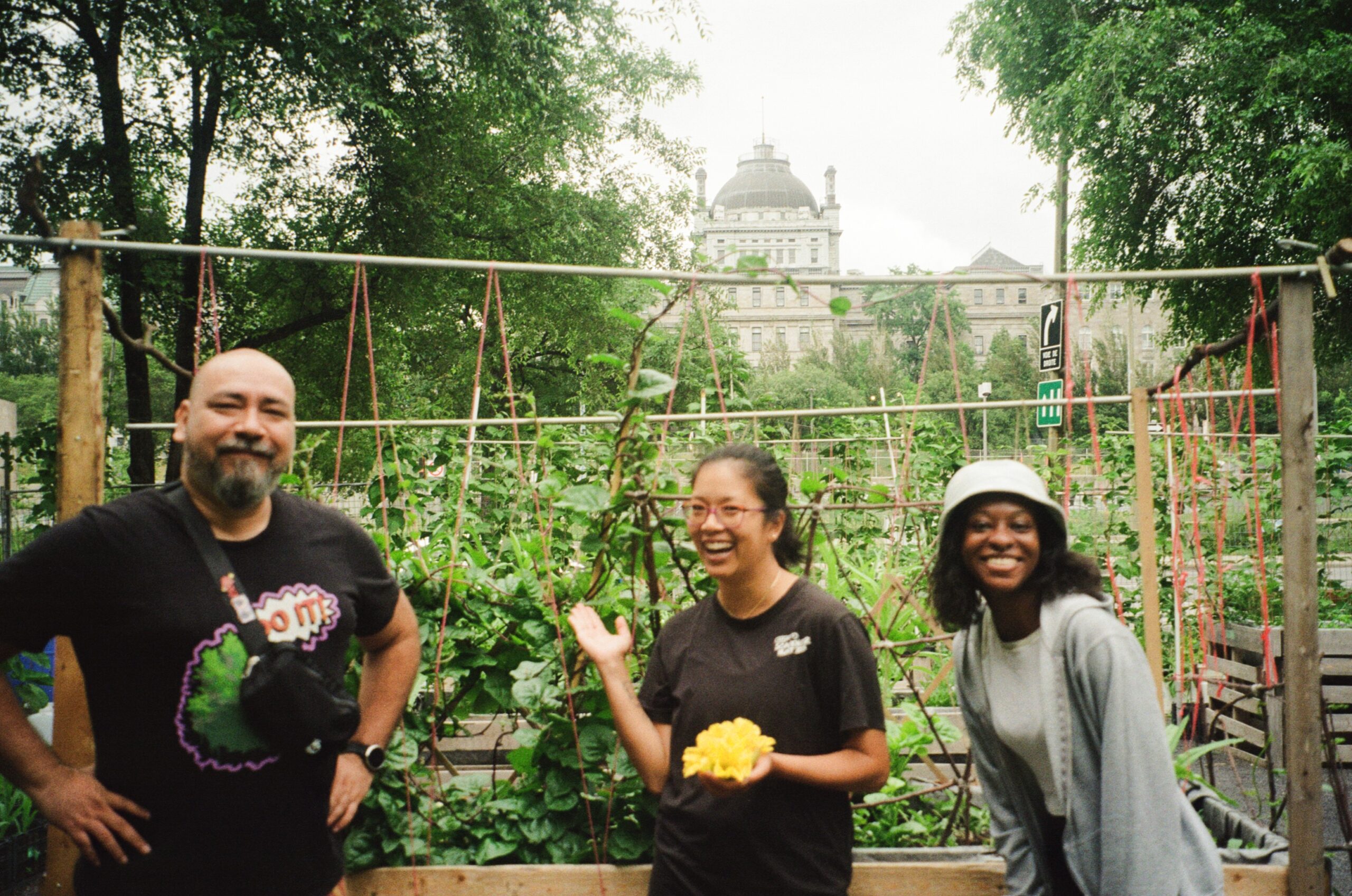Three individuals are standing in a community garden. A man with a beard is smiling with his hands on his hips. A woman in a black shirt holds a yellow flower, gesturing joyfully. To her right, a woman in a light gray sweater and hat smiles. Lush green plants and a building are visible in the background, with trees framing the scene.