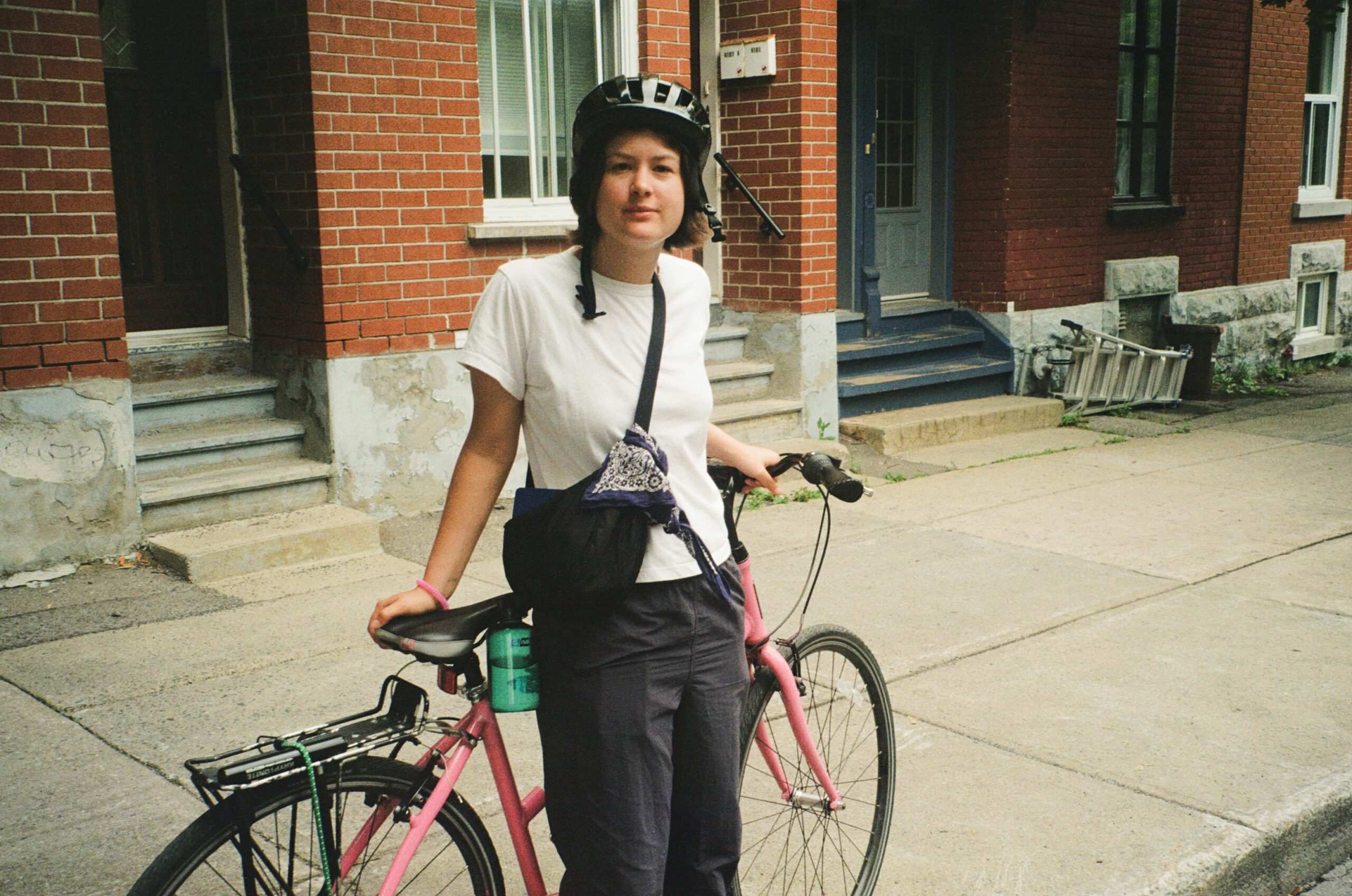 A person wearing a black helmet and a white t-shirt stands beside a pink bicycle on a sidewalk. They have a black bag slung across their shoulder and a green water bottle attached to the bike. The background features a red brick building with steps leading to a doorway.