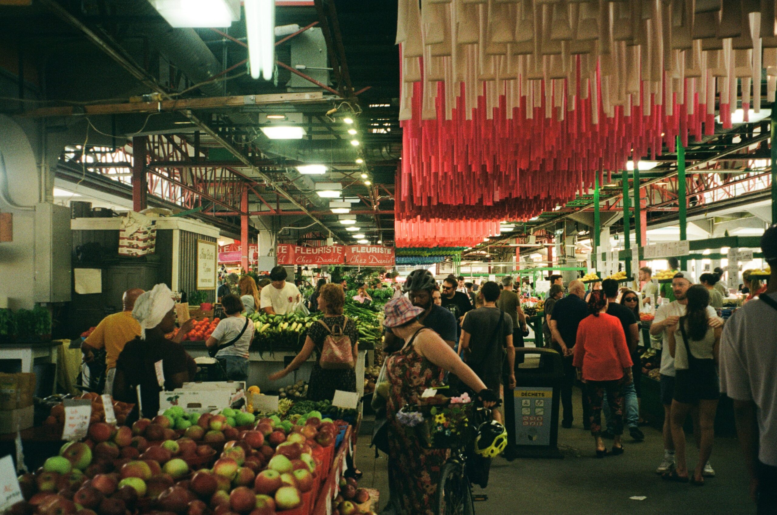 A busy market scene filled with people shopping for fresh produce. Stalls display a variety of fruits and vegetables, prominently featuring apples in baskets. Bright red decorations hang from the ceiling, adding vibrancy to the atmosphere. Shoppers include individuals with bicycles and groups engaging in conversation. Signs in the background hint at various vendors and offerings, creating a lively and colorful marketplace environment.