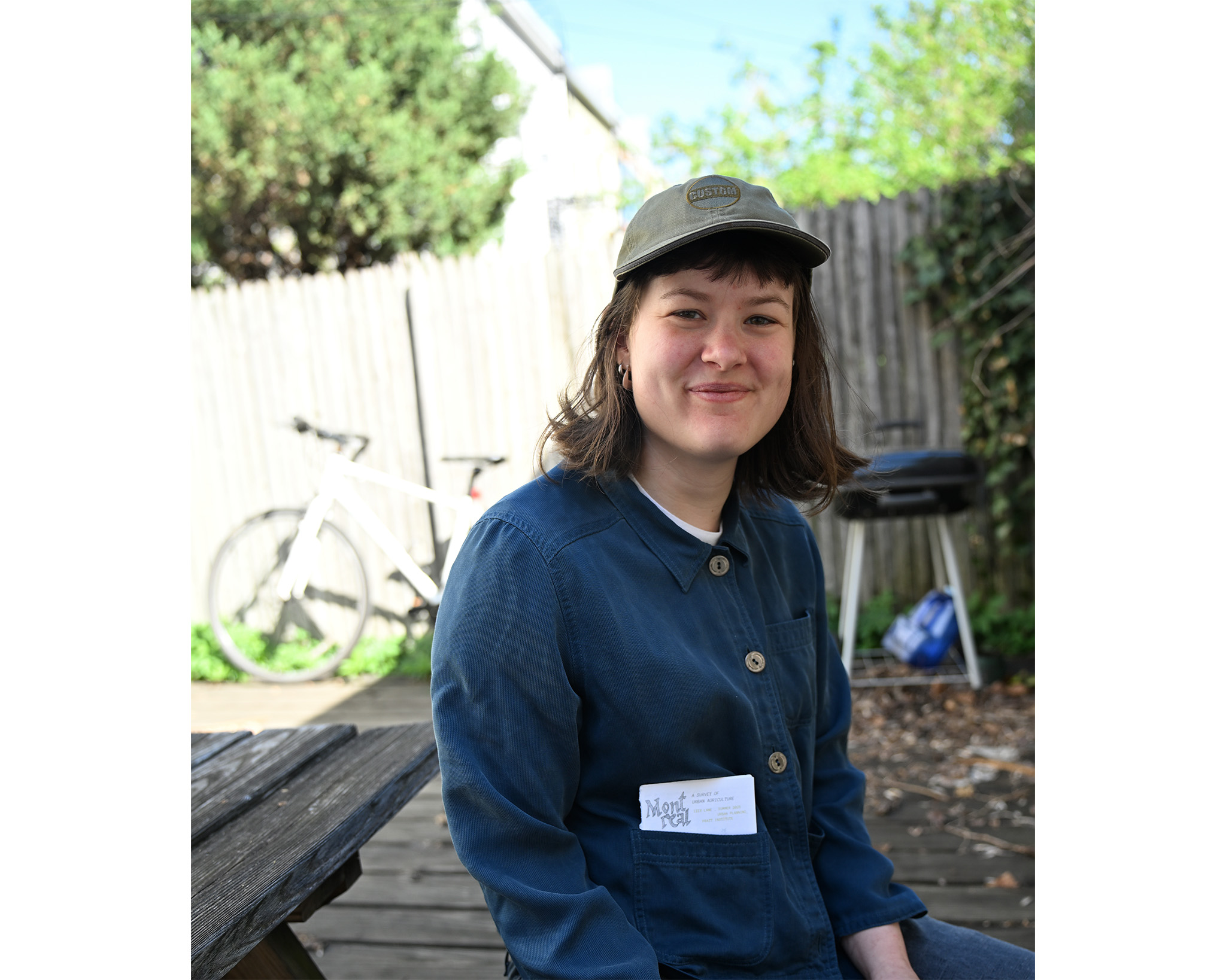 A young person with shoulder-length hair is sitting outdoors, smiling at the camera. They are wearing a blue jacket and a green cap. A name tag is visible on their jacket. In the background, there is a white bicycle and a grill. The setting appears to be a garden or backyard with wooden decking.