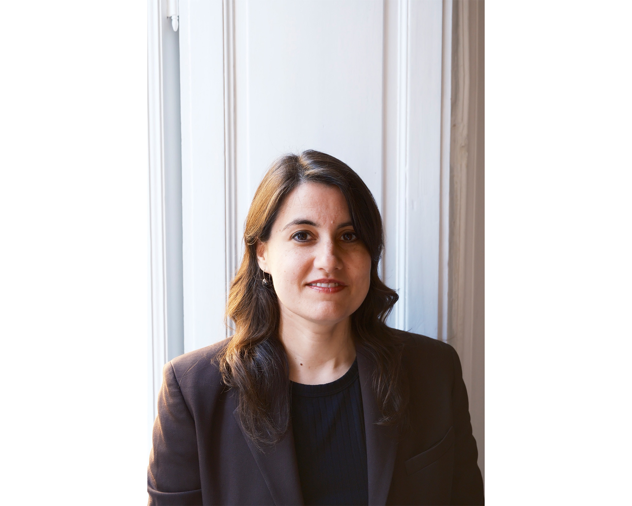 A woman with shoulder-length brown hair is standing near a window with natural light illuminating her face. She is wearing a dark blazer over a black top, smiling softly and looking directly at the camera. The background is a light-colored wall with a window frame.