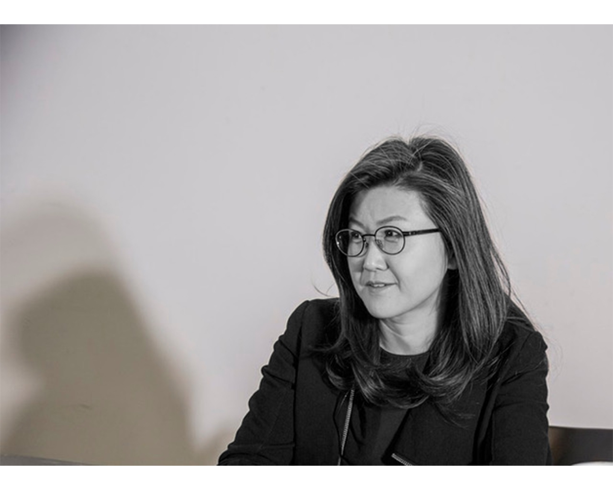 A woman with long, dark hair is wearing glasses and a black blazer, looking to her right. The background is a light color, creating a soft contrast. The lighting is soft and creates subtle shadows.