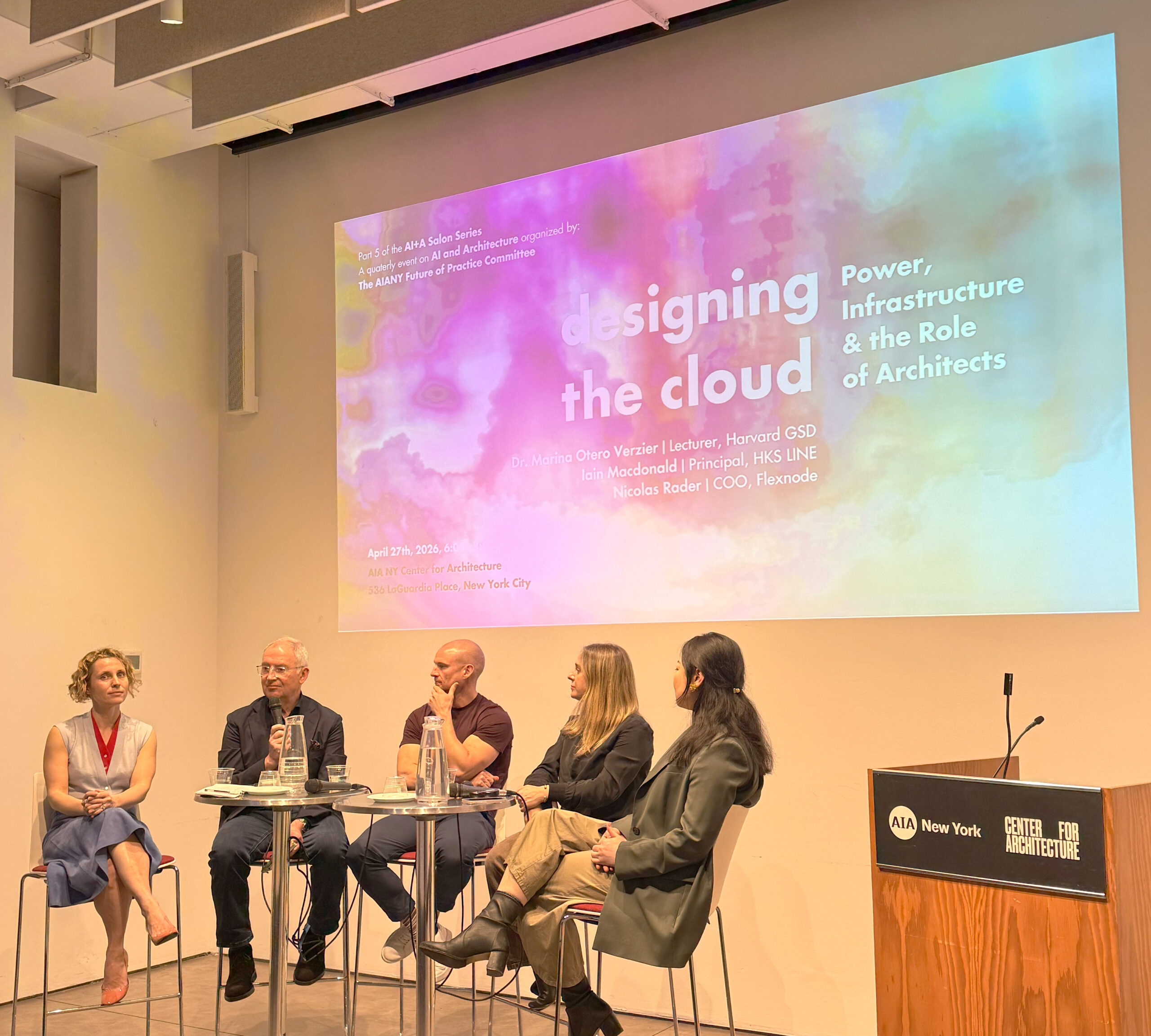 A panel discussion taking place at the AIA Center for Architecture in New York City. Five participants are seated on stools around a table, discussing the topic "designing the cloud." The background features a presentation slide with colorful graphics and text, outlining details of the event, including speakers' names and the date, April 27, 2020. A speaker’s podium is visible on the right side of the image.