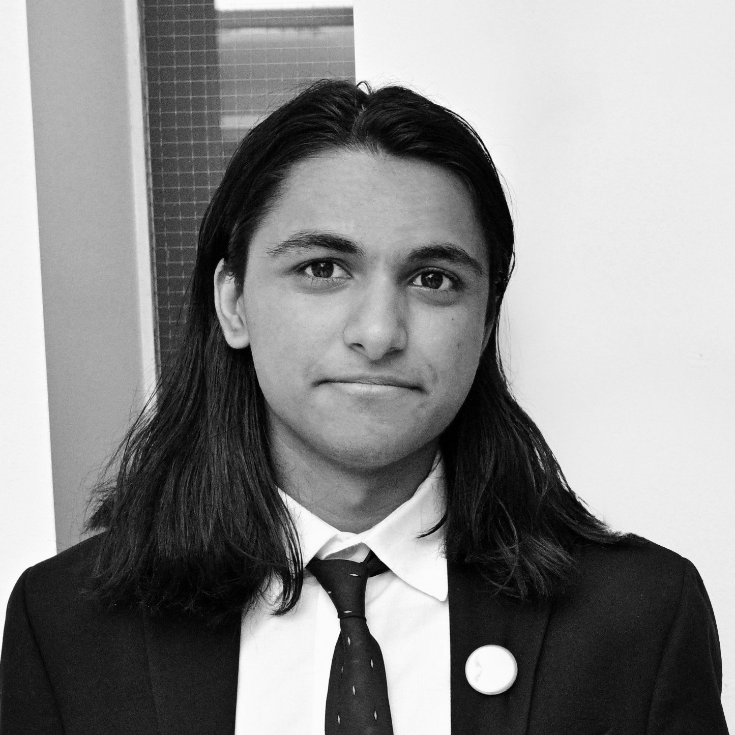 A young person with long black hair is wearing a black suit and a white shirt with a tie. They have a button on their lapel and are looking directly at the camera with a neutral expression. The background features a light wall and a tiled surface. The photo is in black and white.