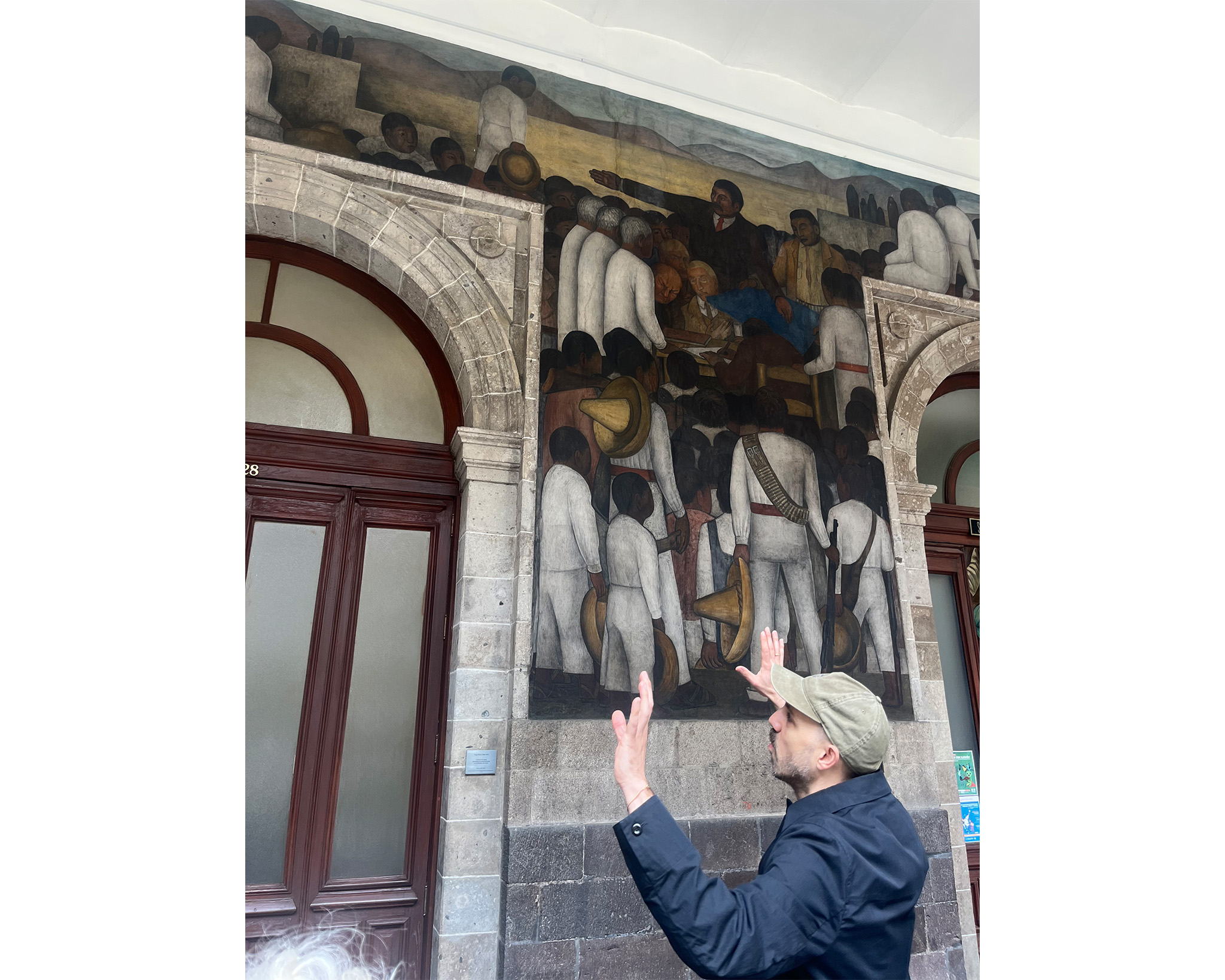 Man poses in raised hand in front of a doorway with a mural by Diego Rivera.