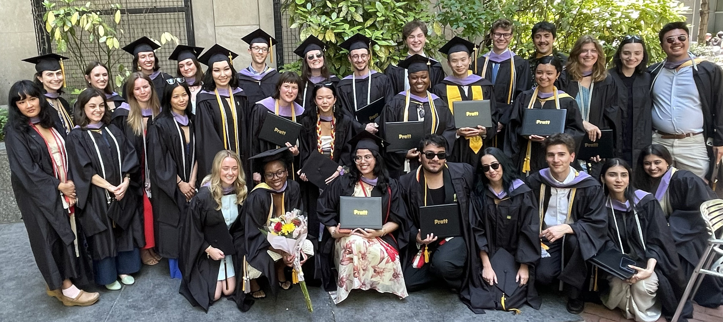 A group of approximately 30 graduates wearing black robes and caps stands outdoors, holding diplomas.