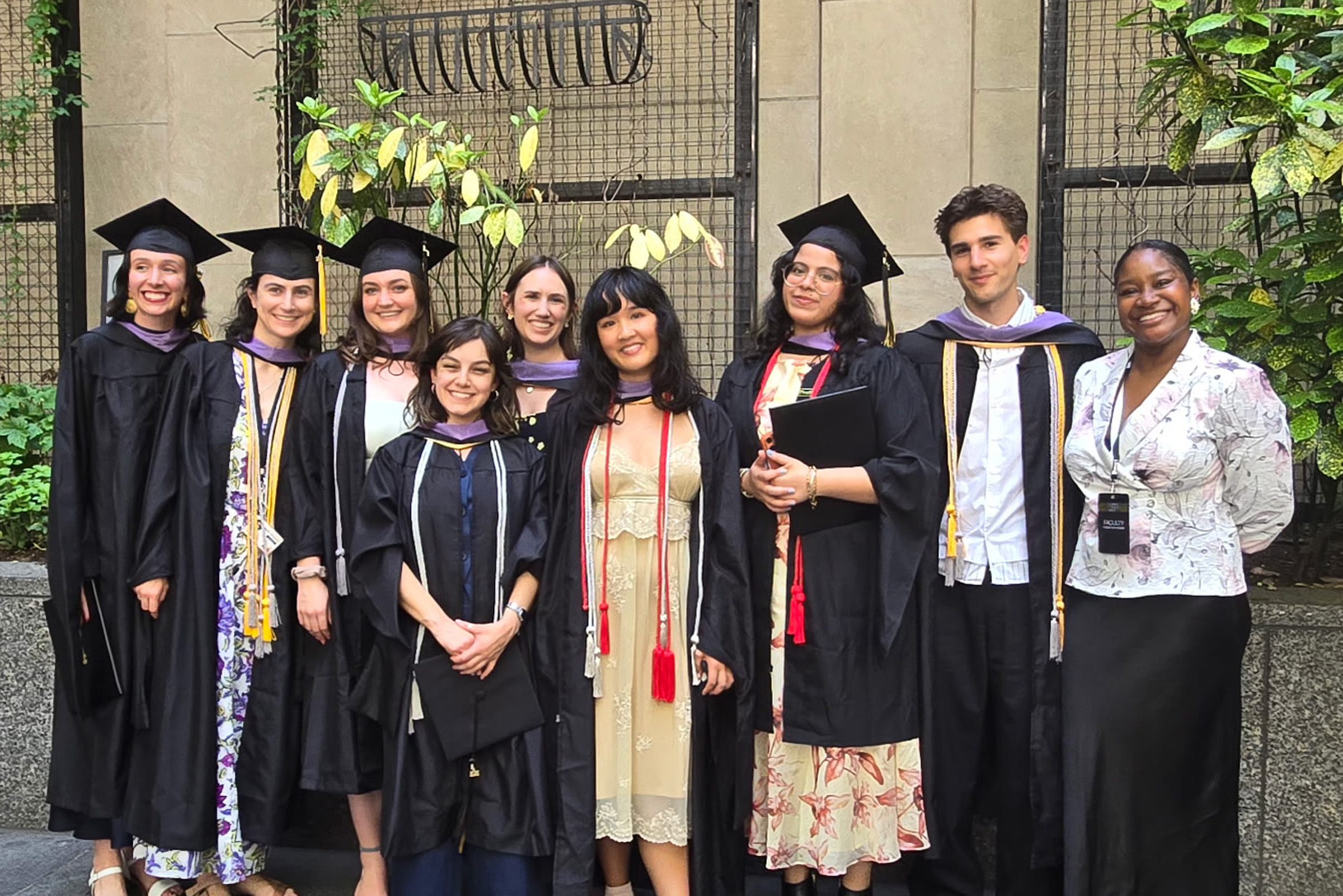 A group of eight students wearing caps and gowns and a woman wearing a floral blouse stand against a background of plants.