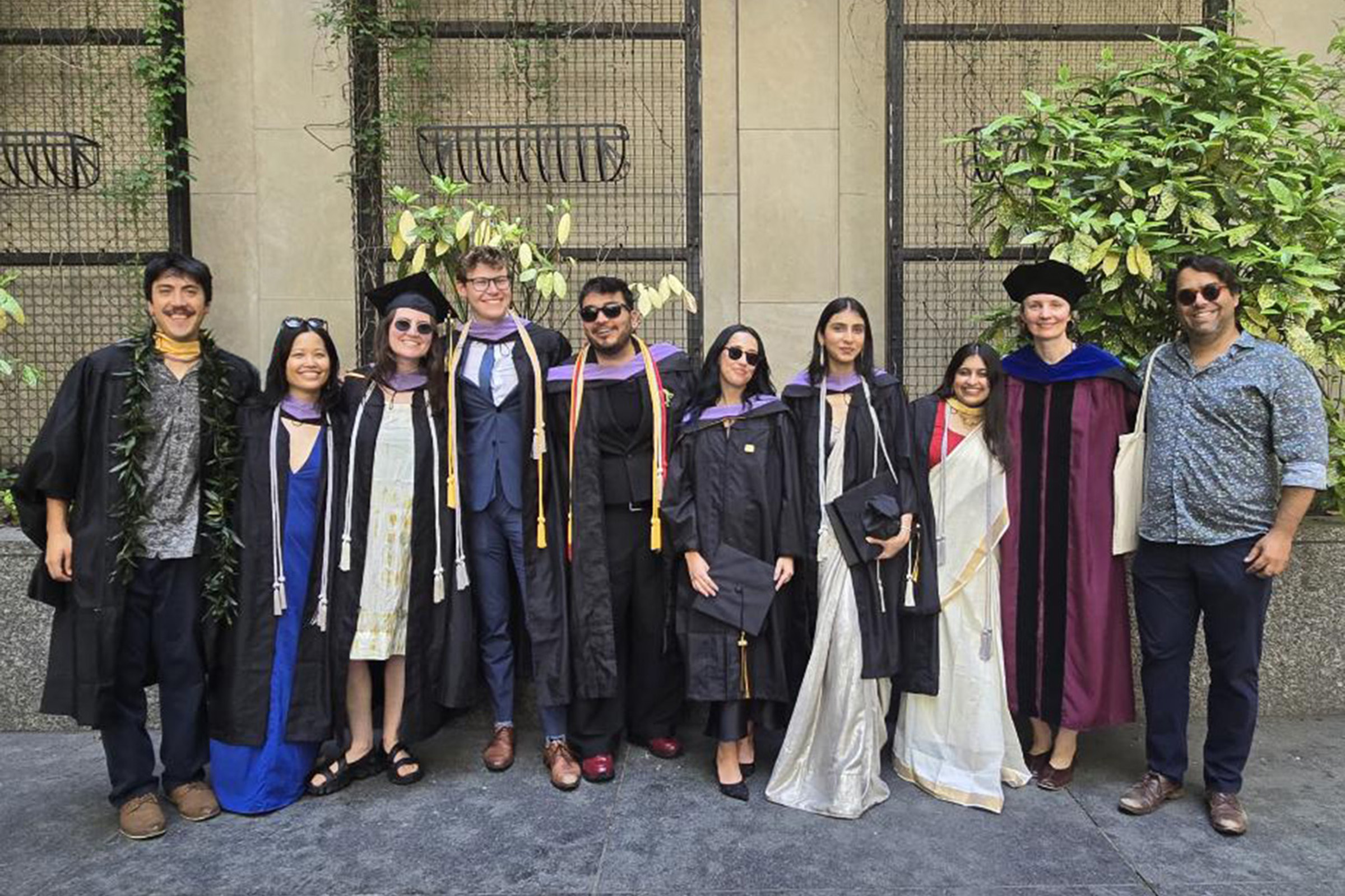 A group of eight students wearing caps and gowns poses with two faculty members against a background of plants.