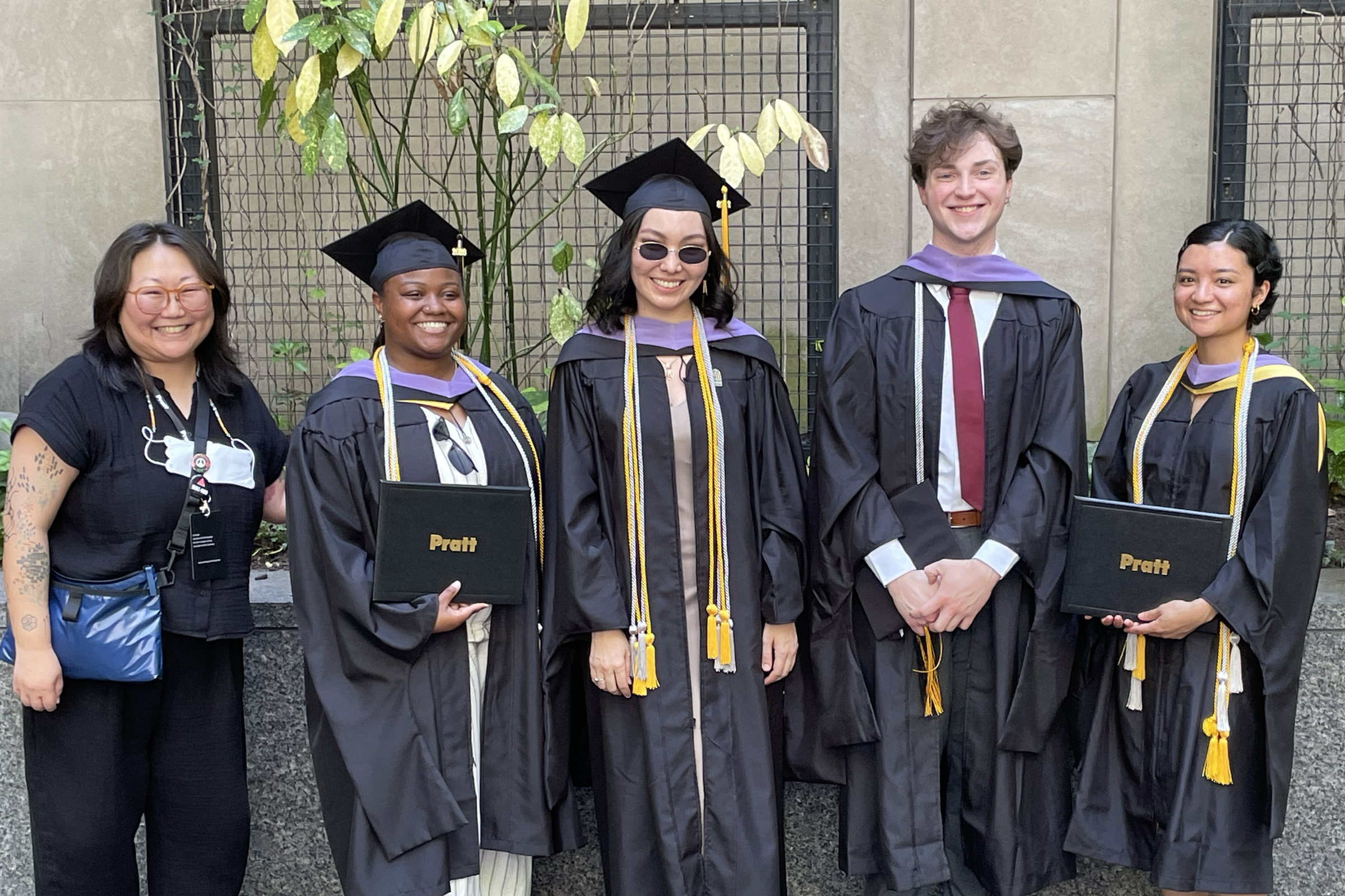 A group of four students wearing caps and gowns poses with their academic director against a background of plants.