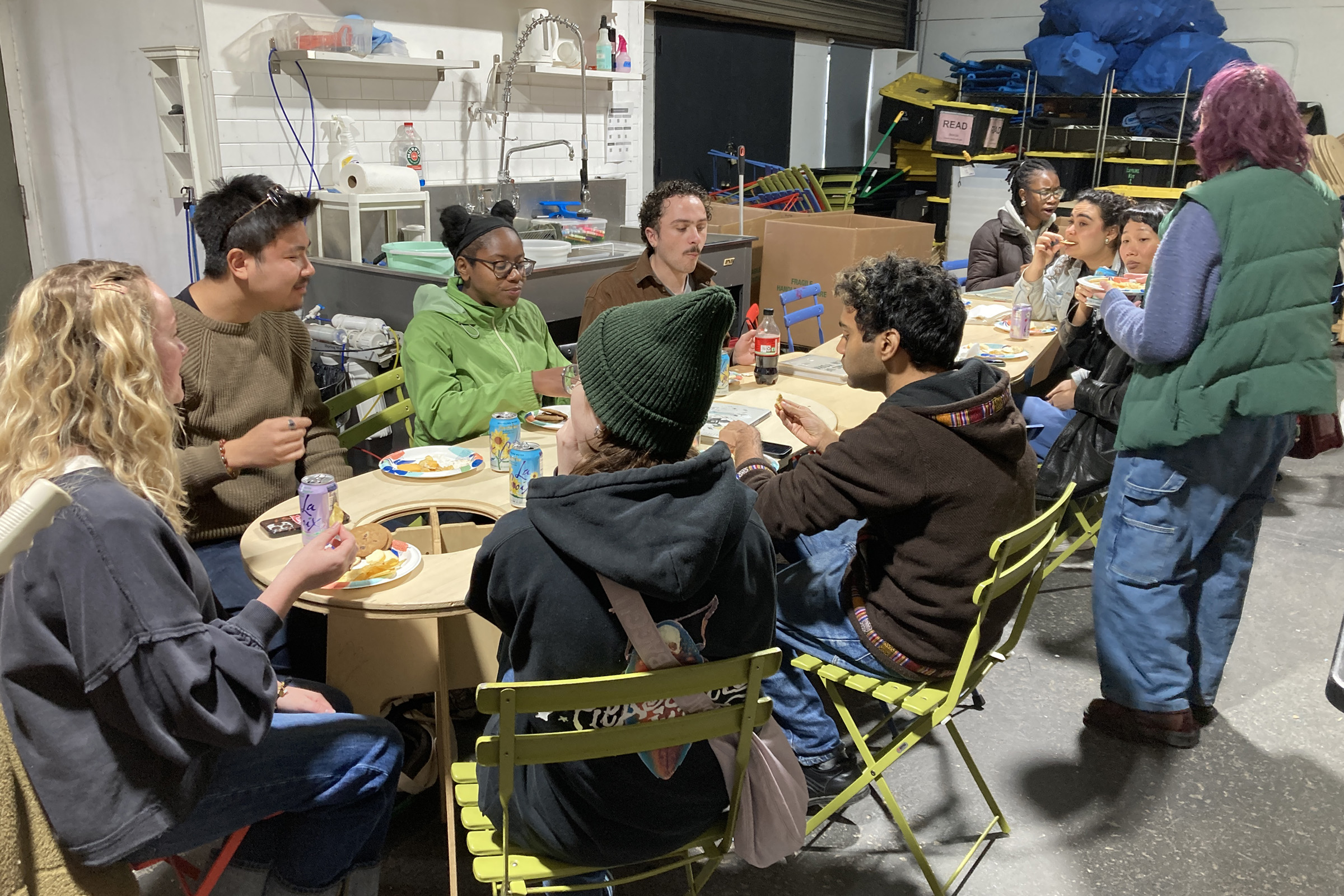 A diverse group of eleven people sits around tables in a casual setting, sharing a meal. Some are enjoying drinks and food, while a woman stands conversing with them. The background includes shelves with various items.