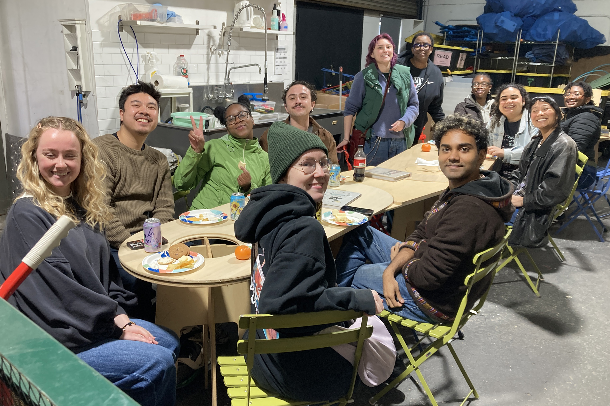 A group of twelve people sit and stand around tables in a casual setting, smiling for the camera. The background includes shelves with various items.