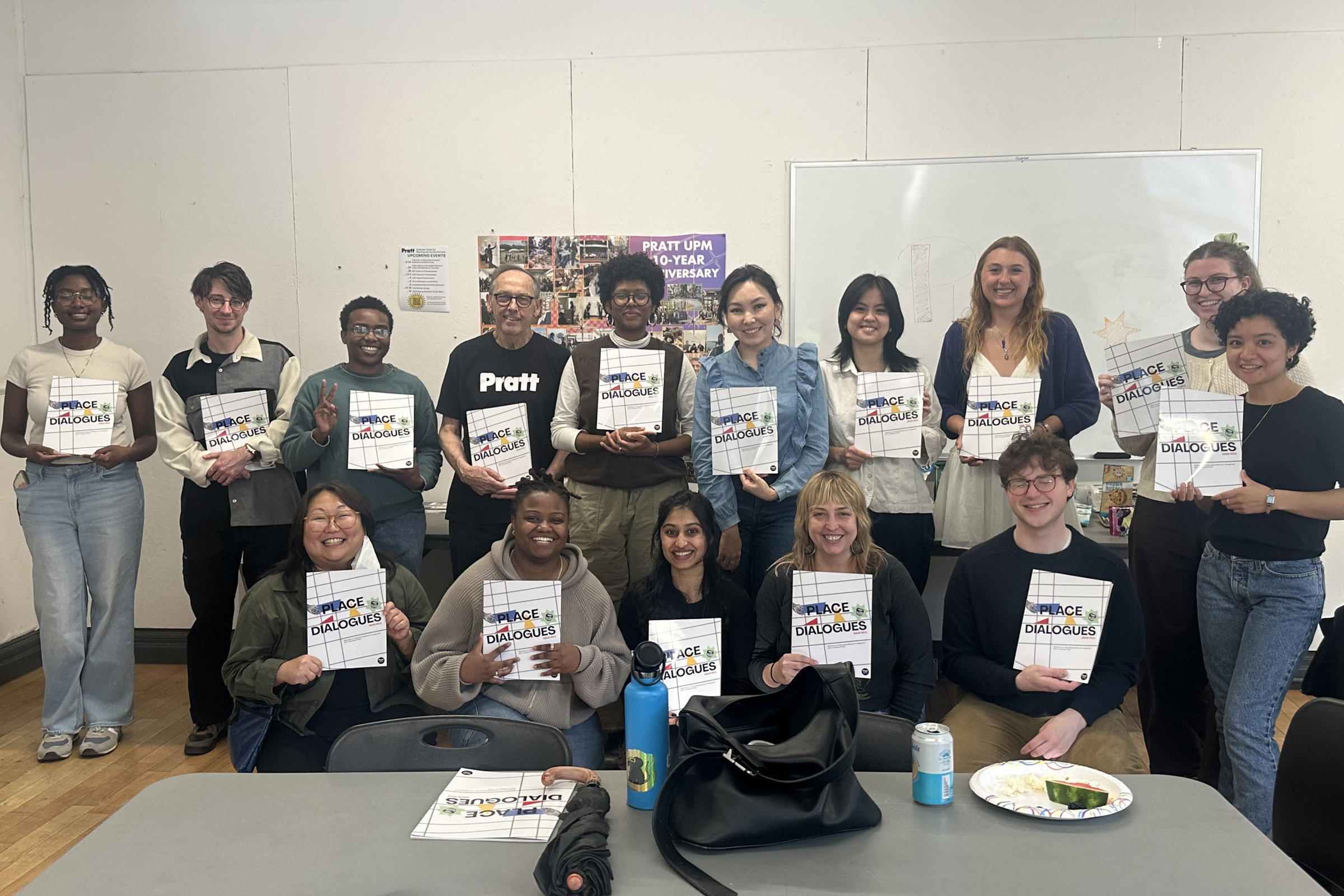 A group of fifteen people holding a publication up, pose together for a photo in a classroom.