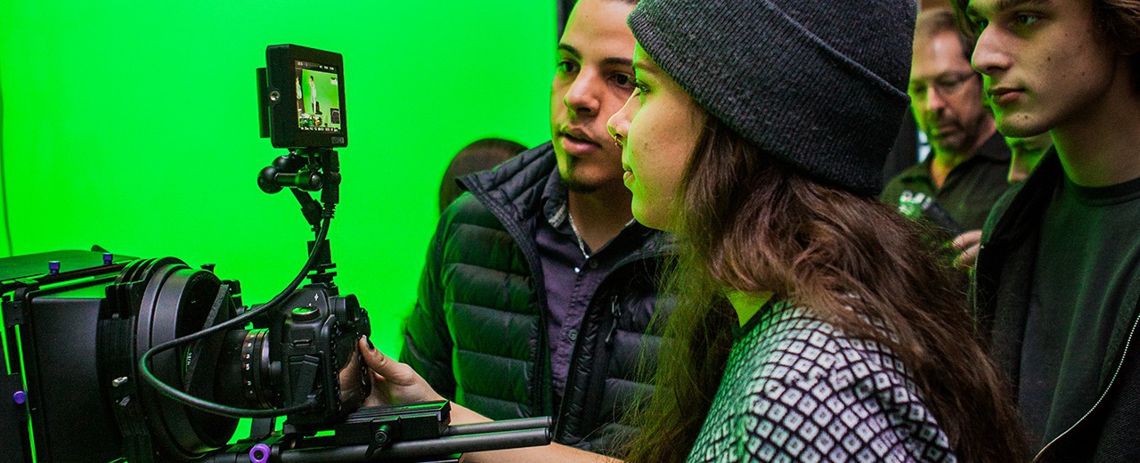 Students gather around a camera as they film a subject with a green screen background.