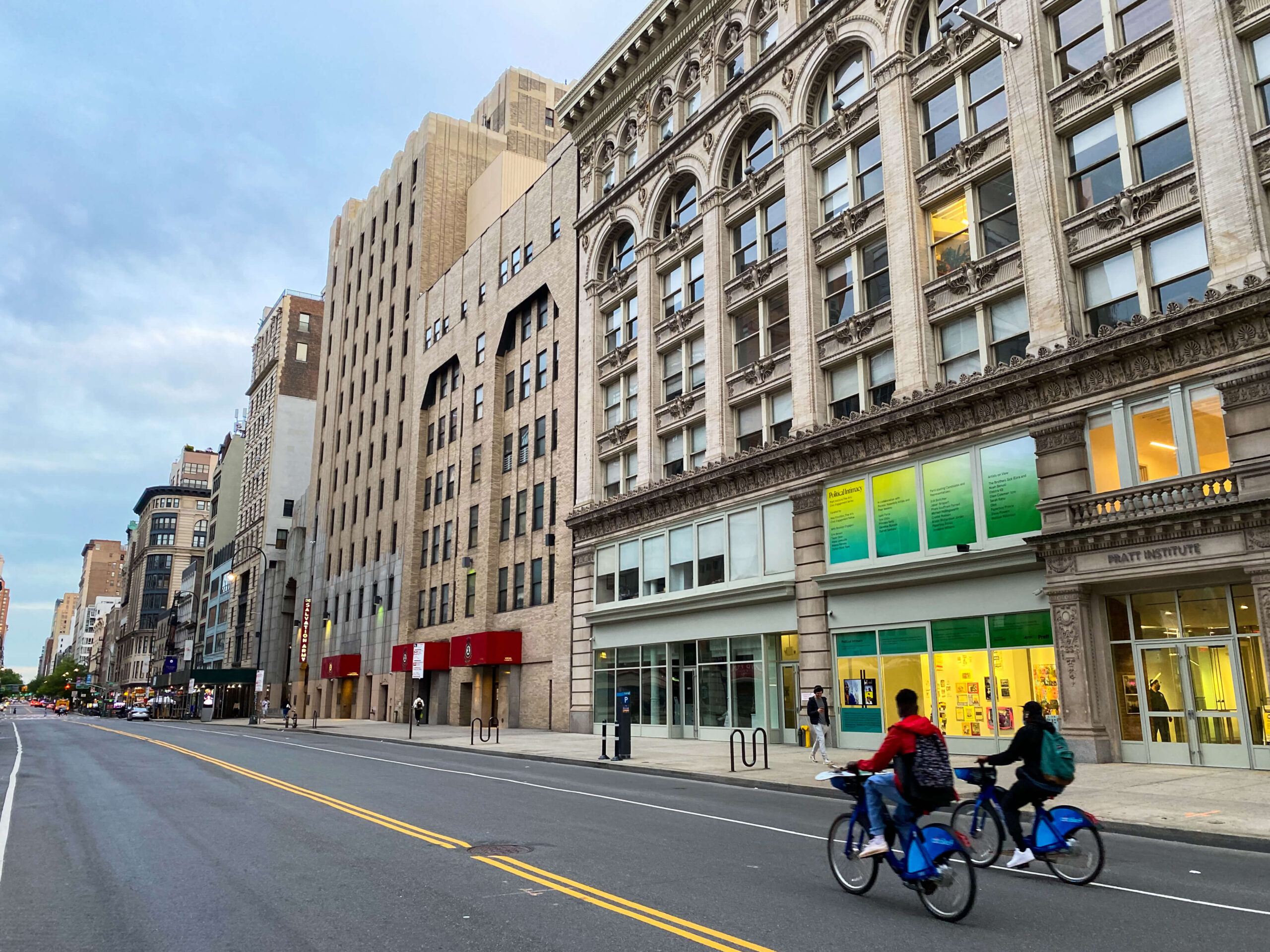 The façade of a grey building with a road directly in front. There are two cyclists going by and people walking on the sidewalk.