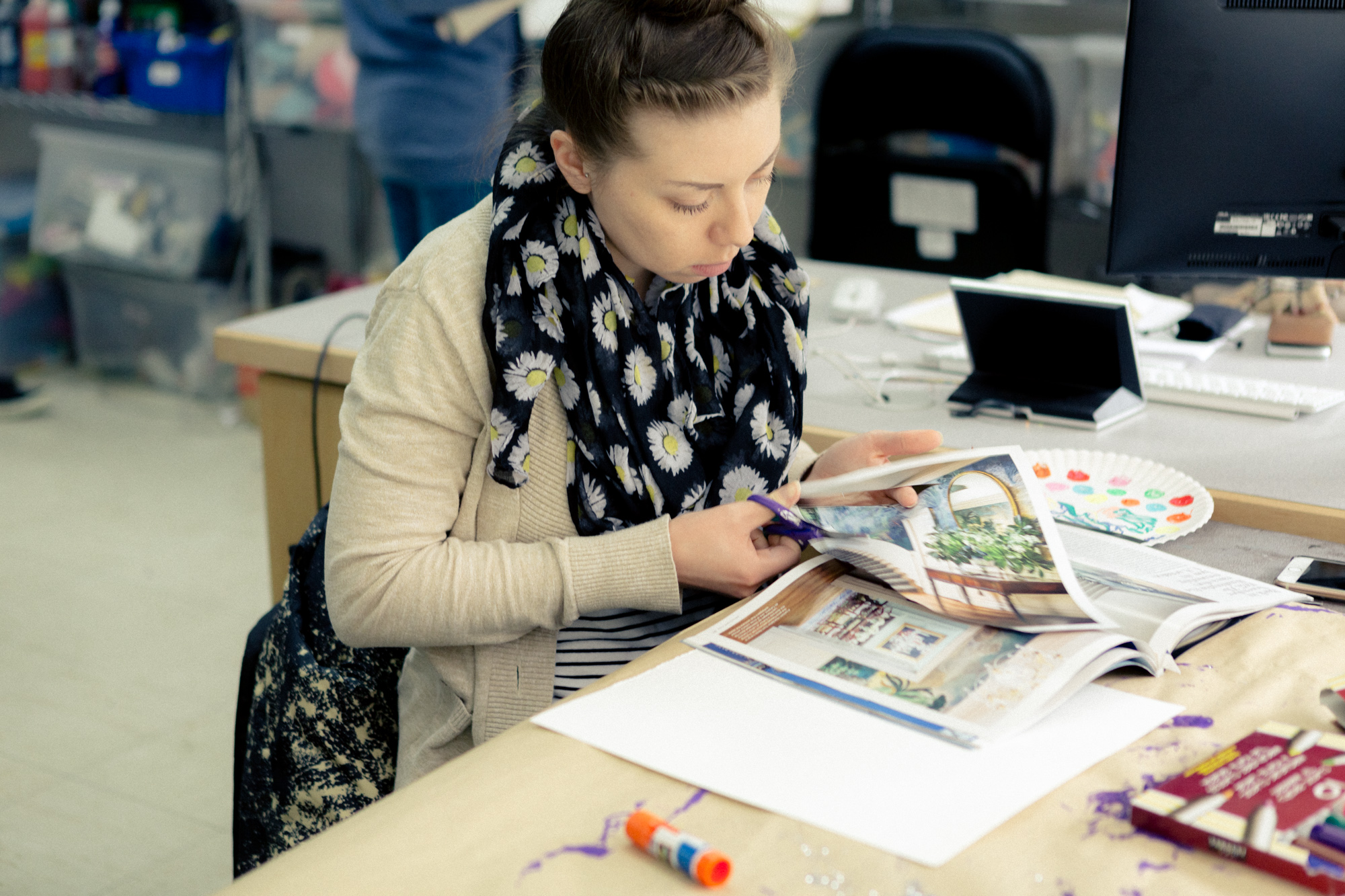 A woman with a scarf covered in sunflowers cuts an image out of a magazine while sitting at a desk.