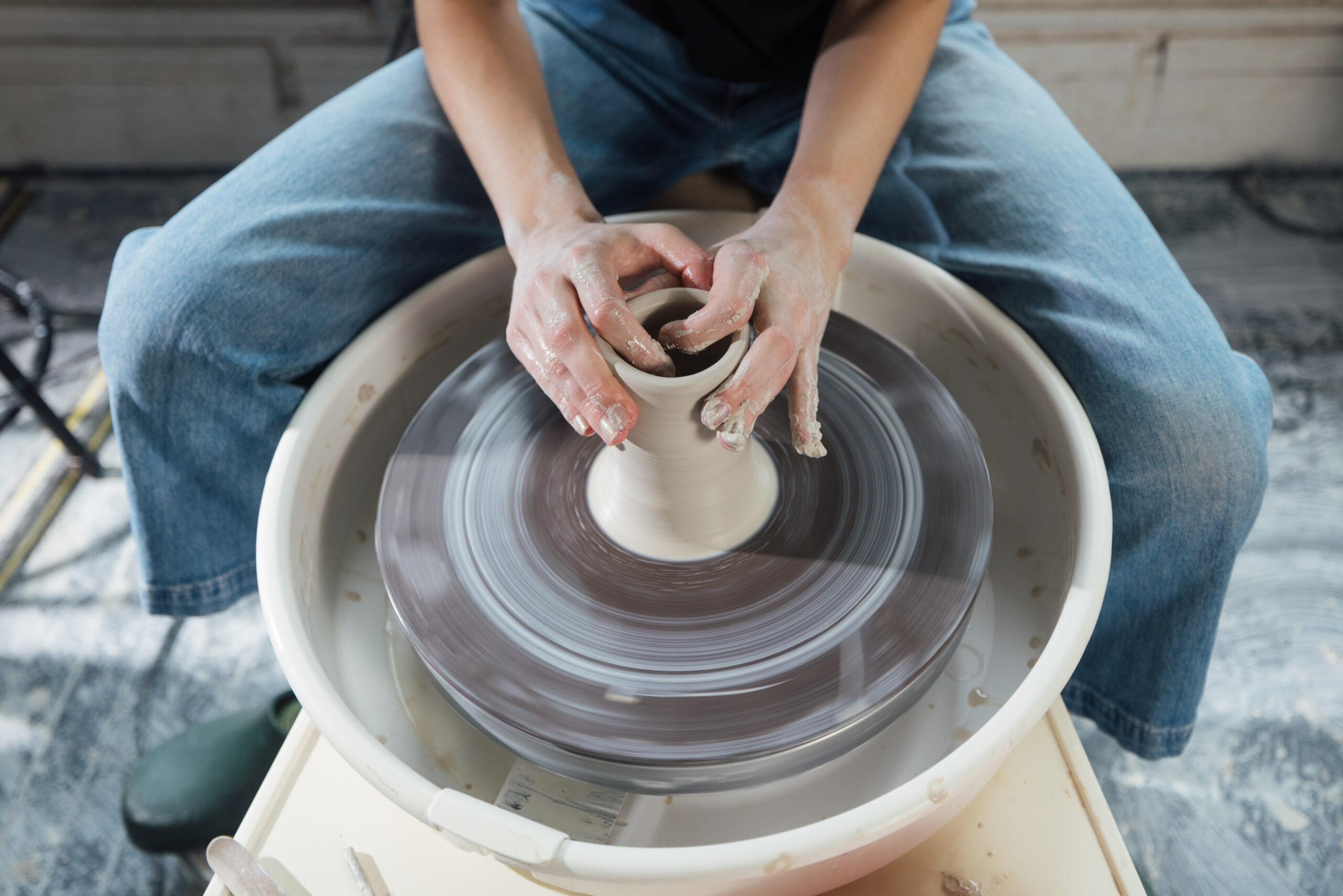 Two hands belonging to a seated person are working with wet clay on a ceramics wheel. The wheel is blurred showing that it is turning. The hands have clay on them from their work. The person is wearing jeans and comfortable shoes. Their torso and above is not visible in the frame.
